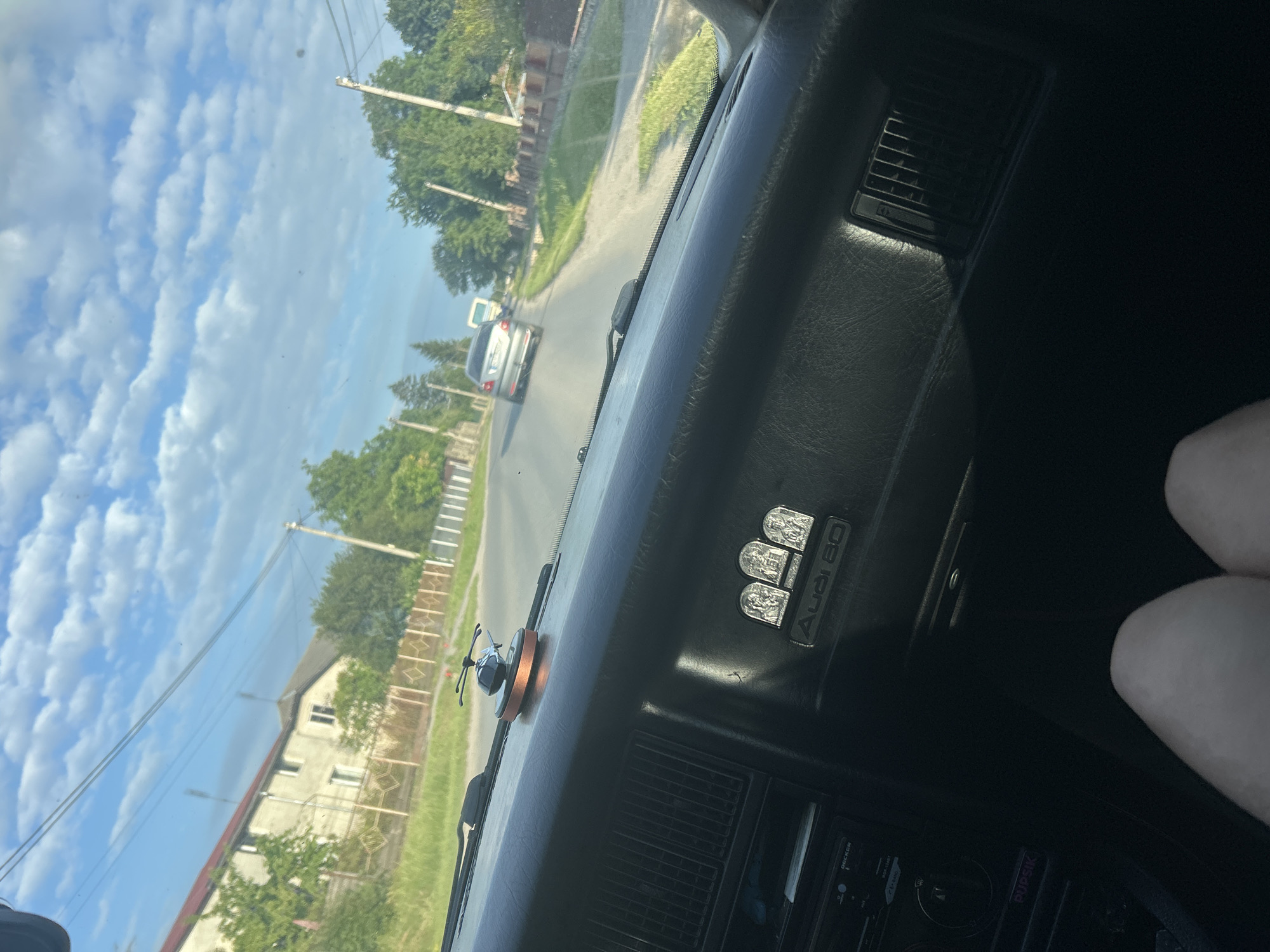 The image provides an interior view from a car driving on a sunny day in Velyki Hai, Ukraine. The dashboard features an "Audi 80" emblem and a small helicopter-shaped air freshener. Outside the car, a paved road is visible with houses and trees lining the street. A silver car is driving ahead on the road. The sky is filled with fluffy white clouds against a clear blue backdrop, suggesting pleasant weather. The time of day appears to be afternoon, given the bright sunlight and the shadows cast. The overall scene depicts a peaceful drive through a residential area.