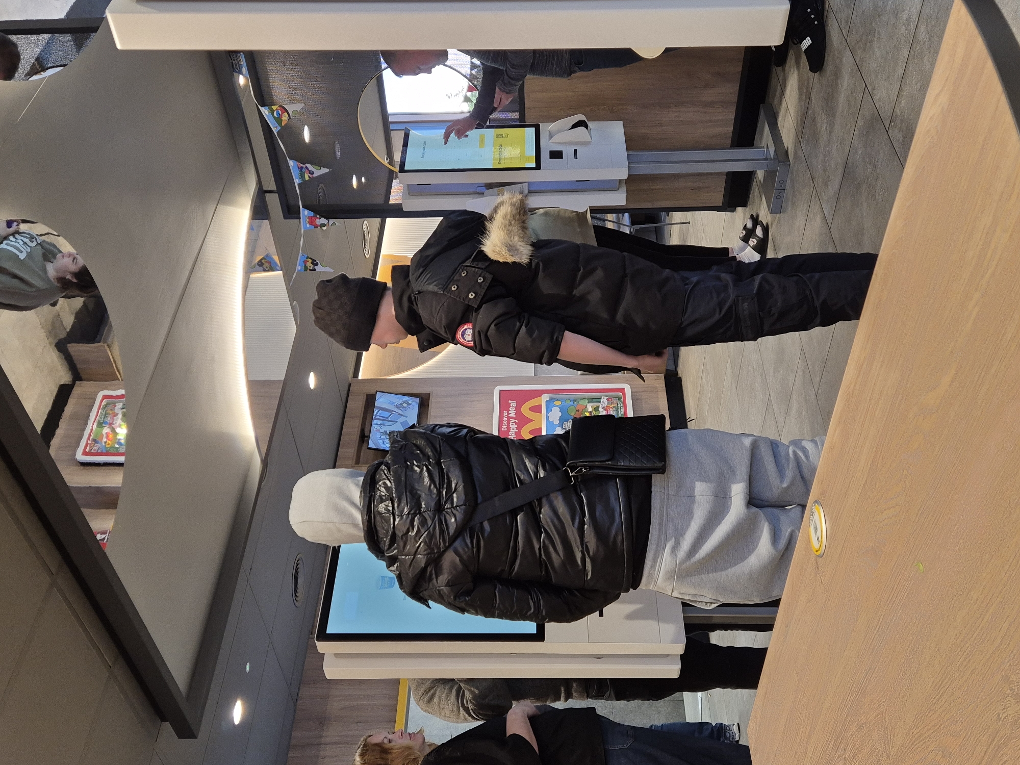 The image depicts the interior of a McDonald's restaurant in West Suffolk, United Kingdom. Two individuals are interacting with self-service kiosks. One person, wearing a black beanie and a black jacket with a fur-lined hood, is looking at the screen of the kiosk. The other individual, wearing a grey hooded sweatshirt and a black puffer jacket with a small black purse slung across their chest, is also interacting with a kiosk. The kiosks are white with touch screen displays. In the background, other patrons are visible, some standing and others reflected in a mirrored surface. The decor includes a banner with cartoon character images, and promotional material featuring "Happy Meal." A wooden table is visible to the side. The lighting is bright, and the overall impression is of a modern fast-food establishment.