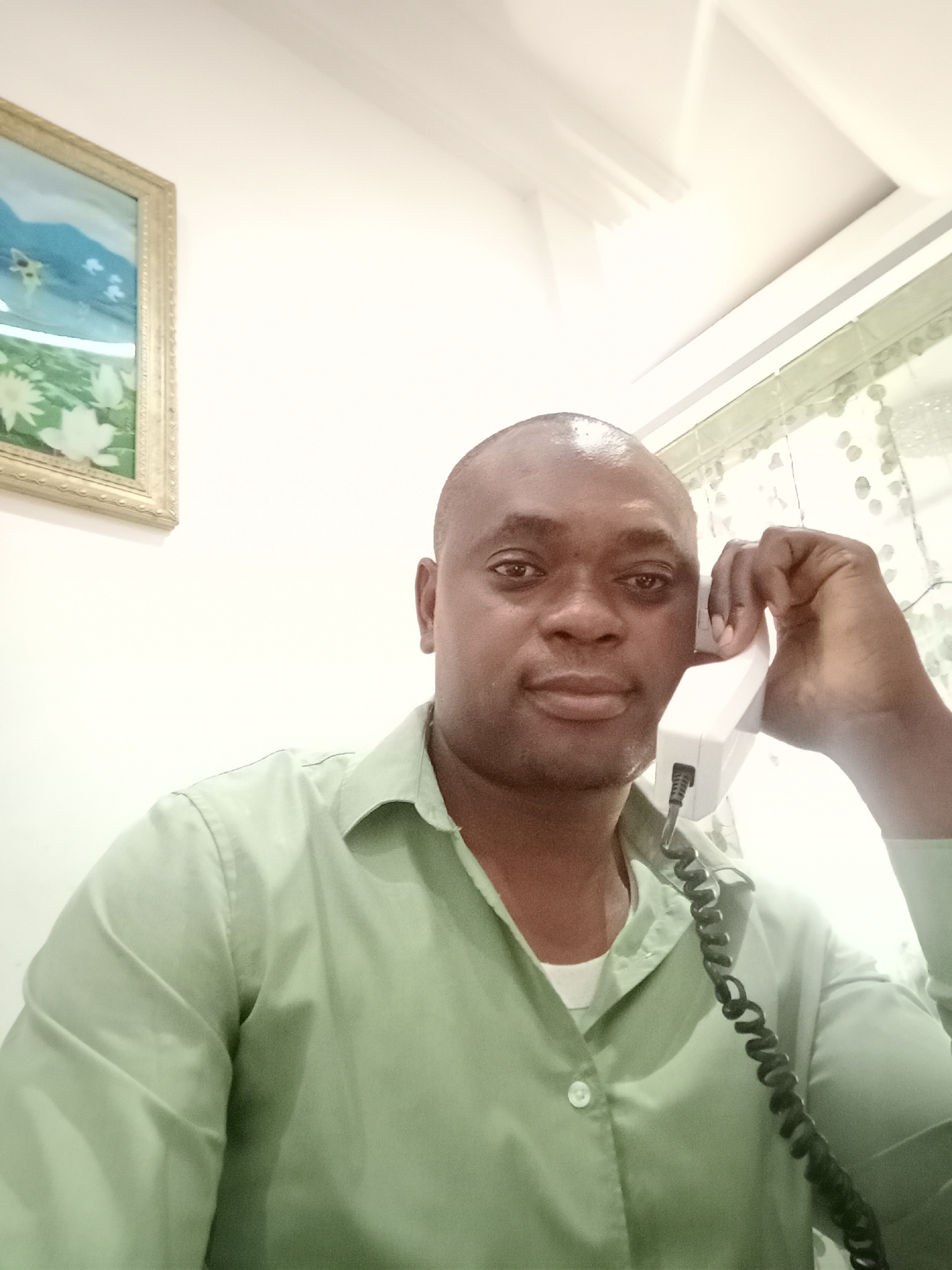 A dark-skinned man with a shaved head is holding a white telephone receiver to his left ear. He is wearing a light green button-down shirt with a white undershirt visible at the collar. A coiled black telephone cord is visible against the shirt. The background includes a white wall with a framed painting on the left and what appears to be a window with a patterned curtain on the right. The location is likely indoors and may be in the city of Gwarinpa, Nigeria, although there are no specific location markers in the image. The man appears to be on a phone call.