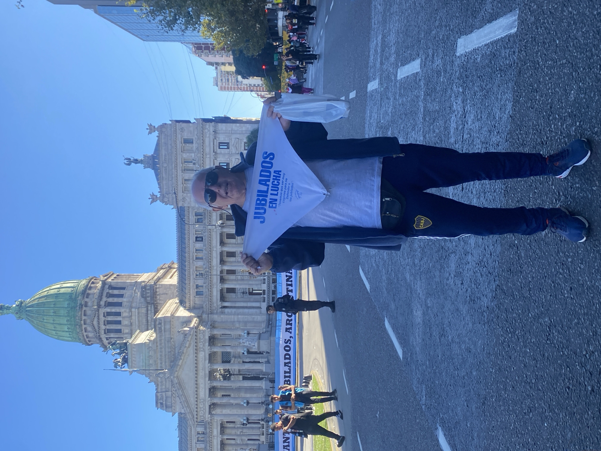 The image captures a man standing on a street in Buenos Aires, Argentina, participating in what appears to be a protest or demonstration. The man, positioned prominently in the foreground, wears sunglasses, a grey t-shirt, a dark jacket, dark sweatpants featuring a yellow patch that resembles the Boca Juniors crest, and blue sneakers. He holds a white, triangular flag or banner with blue text.
The visible text on his banner reads: "JUBILADOS EN LUCHA" (Retirees in Struggle), with smaller text beneath stating: "Tenemos que ser muy cagones para no Defender a los jubilados" (We have to be very cowardly not to defend retirees). Around his waist, he wears a dark fanny pack.
The background prominently features the majestic Palacio del Congreso de la Nación Argentina (National Congress Palace), with its iconic green dome and intricate neoclassical architecture. The setting is an open street under a clear, bright blue sky, indicating sunny daytime conditions. Further down the street, other people are visible, suggesting a larger gathering. A long banner is stretched across the road in the distance, partially obscuring the word but clearly showing "...BILADOS, ARGENTINA," which likely reads "JUBILADOS, ARGENTINA" (Retirees, Argentina). White road markings are visible on the asphalt surface. The scene strongly conveys a public demonstration advocating for the rights of retirees.