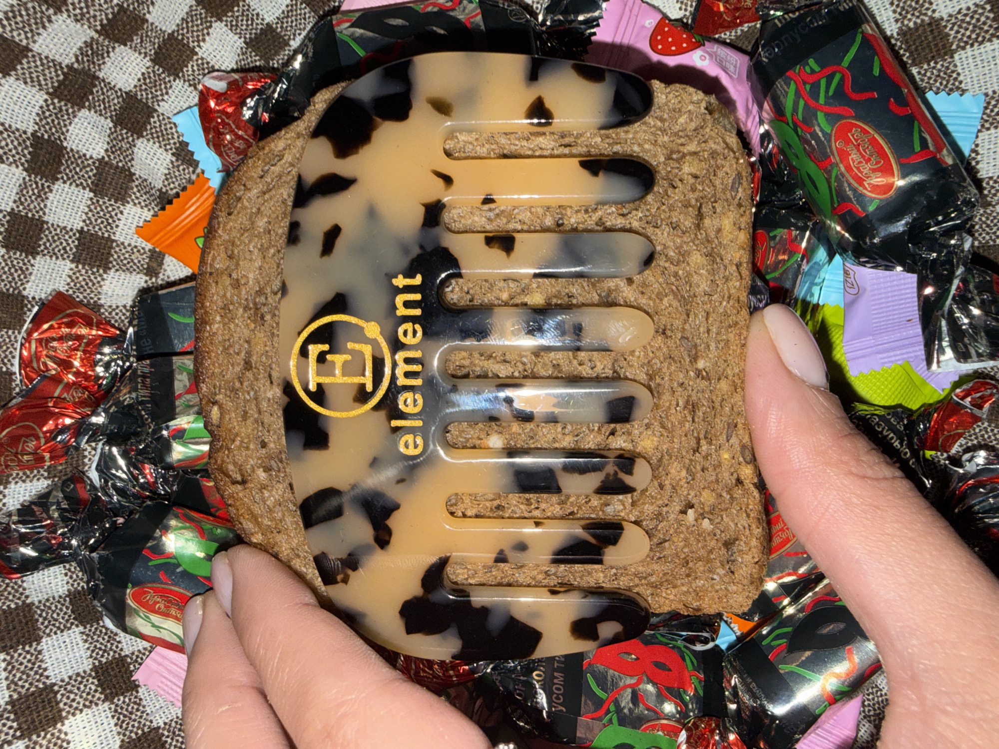 The image shows a brown and black patterned hair comb, labeled "Element", being held over a piece of rye bread. The background is a mix of Russian candies and a brown and white checkered cloth. The candies are wrapped in various colors, including red, black, pink, orange, and blue. Some candies feature designs such as masquerade masks and strawberries. A partial view of a person's hand and fingers is visible holding the comb in place. The location is not specified or identifiable from the image.