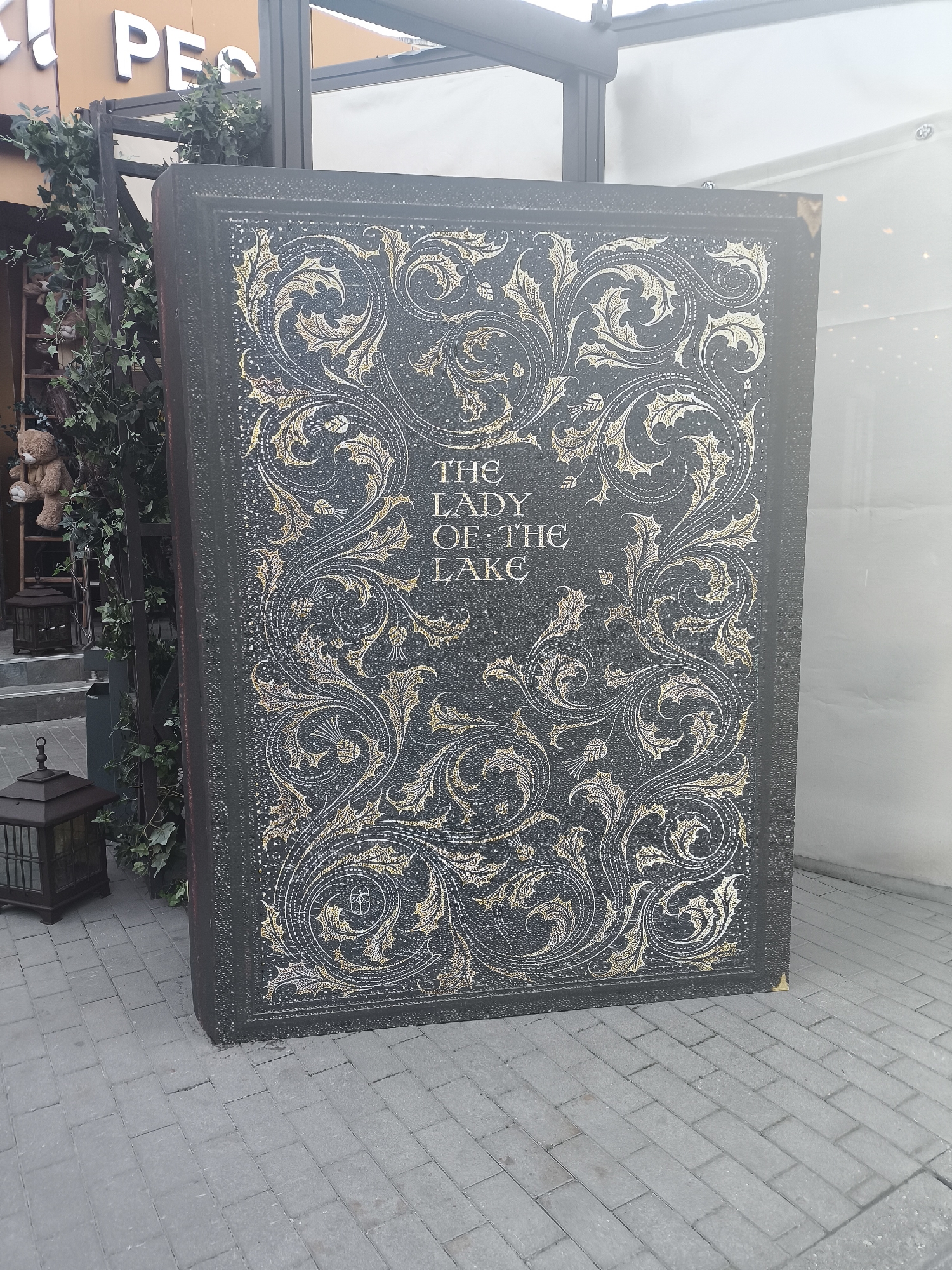 Here is a detailed description of the image:
**Location**:
The scene appears to be set outdoors, possibly near a commercial establishment or a tourist attraction in an urban area.
**Objects and Subjects**:
* **Large Book Prop:** A massive, rectangular book prop with a dark blue or black cover dominates the frame. Its cover is adorned with elaborate, swirling golden designs. The title "THE LADY OF THE LAKE" is prominently displayed in the center of the cover.
* **Building/Structure:** The book is positioned next to a building or structure. The structure has a facade with a window or transparent panel.
* **Vegetation:** Lush greenery, possibly ivy, is seen on the left side of the frame, wrapping around a railing or other structural element.
* **Lantern:** A decorative, vintage-style lantern is visible in the left foreground.
* **Paved Surface:** The book and the lantern are resting on a paved surface, which is likely a sidewalk or pedestrian area. The paving is made of rectangular bricks.
* **Teddy Bear:** In the background, on some shelving or a staircase, there is a teddy bear.
**Actions and Interactions**:
There are no visible people or actions directly depicted in the image. The scene is static, with the primary focus on the book prop and its immediate surroundings.
**Environment**:
The environment seems to be a mix of decorative and functional urban elements. The presence of greenery suggests an attempt to create a more inviting or picturesque atmosphere. The overall lighting is bright, casting subtle shadows around the objects.