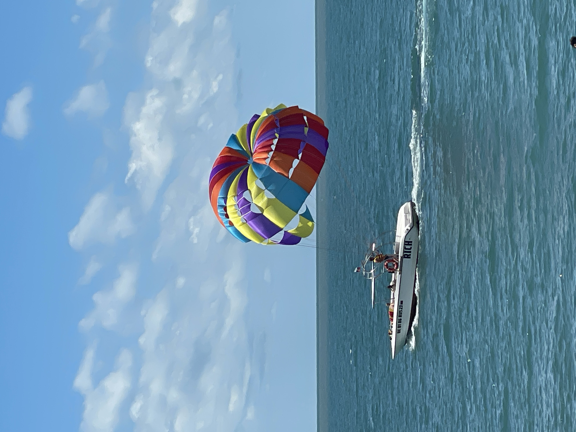 The image depicts a bright, sunny day at sea, likely in the city of Sochi, Russia, given the context provided. A white motorboat, identified by the name "RICH" on its stern, is in motion, leaving a white wake on the blue-green ocean. The boat is carrying several individuals, including a person in a yellow vest who appears to be supervising or operating the boat. Attached to the boat by a tow rope is a large, colorful parasail, featuring vibrant stripes of red, orange, yellow, purple, and blue. The parasail is inflated and soaring in the sky, its multicolored canopy dominating the upper right portion of the image. The background consists of a clear blue sky dotted with fluffy white clouds, meeting the horizon line of the vast ocean. The scene captures a recreational activity, likely parasailing, taking place on a beautiful day.