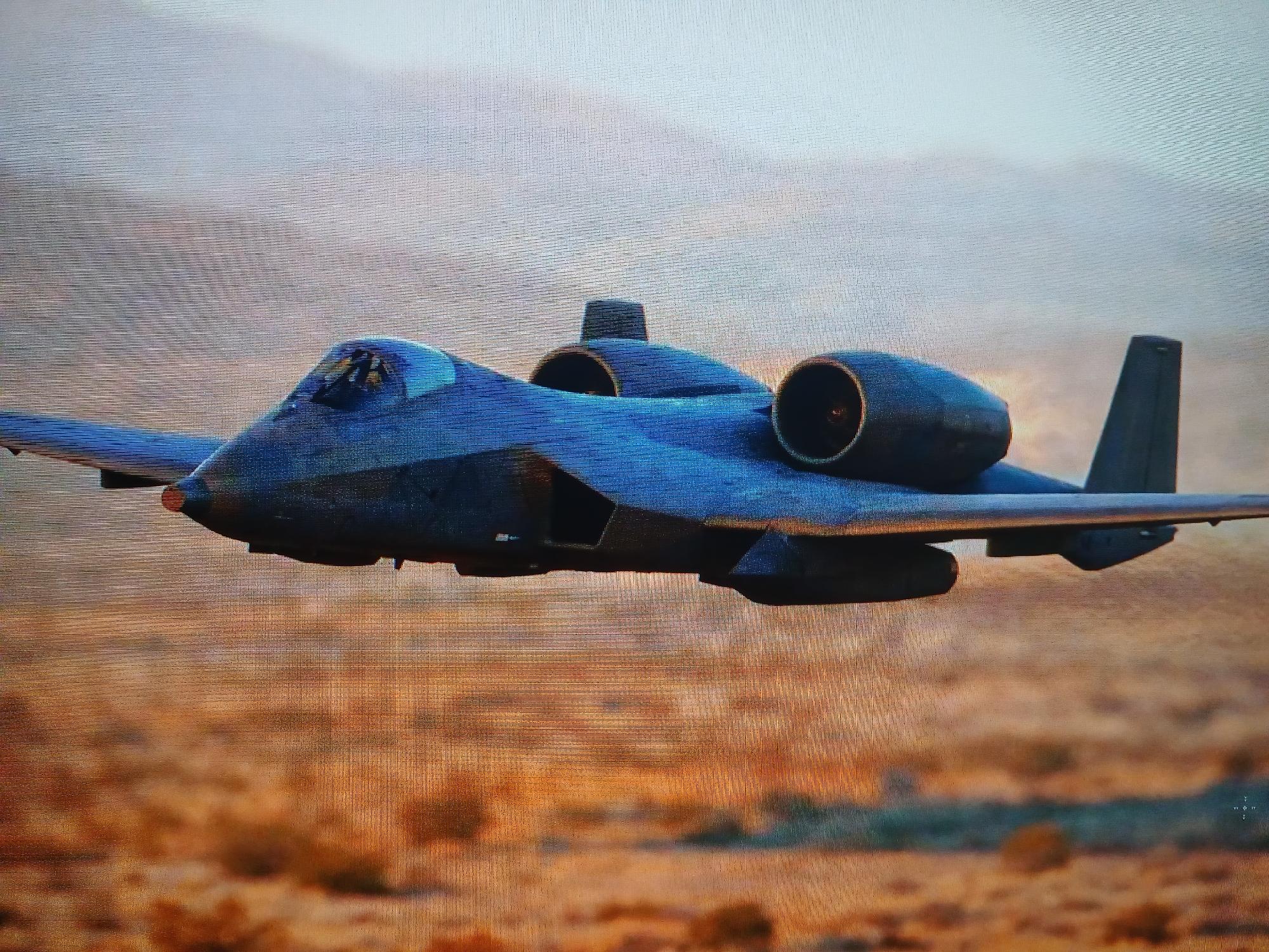 A military aircraft is in flight over a desert-like terrain. The aircraft appears to be a stylized, potentially fictional or modified version of an A-10 Thunderbolt II, featuring twin engines mounted on the fuselage's top, a straight wing design, and a single vertical stabilizer. The environment below is a low-lying, arid landscape with hues of brown and orange. The sky is overcast or hazy. The location is not Kamianske, Ukraine.