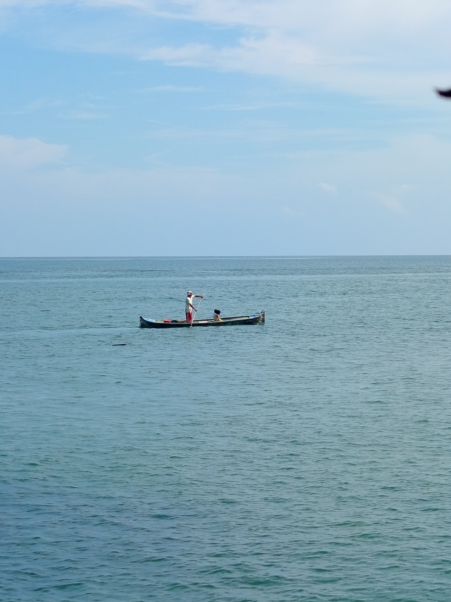 Two people are in a long, narrow boat on the ocean. The person in the back, dressed in a white shirt and red pants, is standing and using a long pole to propel the boat through the water. The person in the front is sitting and appears to be a child. The boat is moving from left to right. The ocean is calm with gentle waves, and the sky is mostly clear with a few wispy clouds. The scene appears to be in a coastal area, possibly near the city of Tolú, Colombia, known for its beaches and ocean activities.