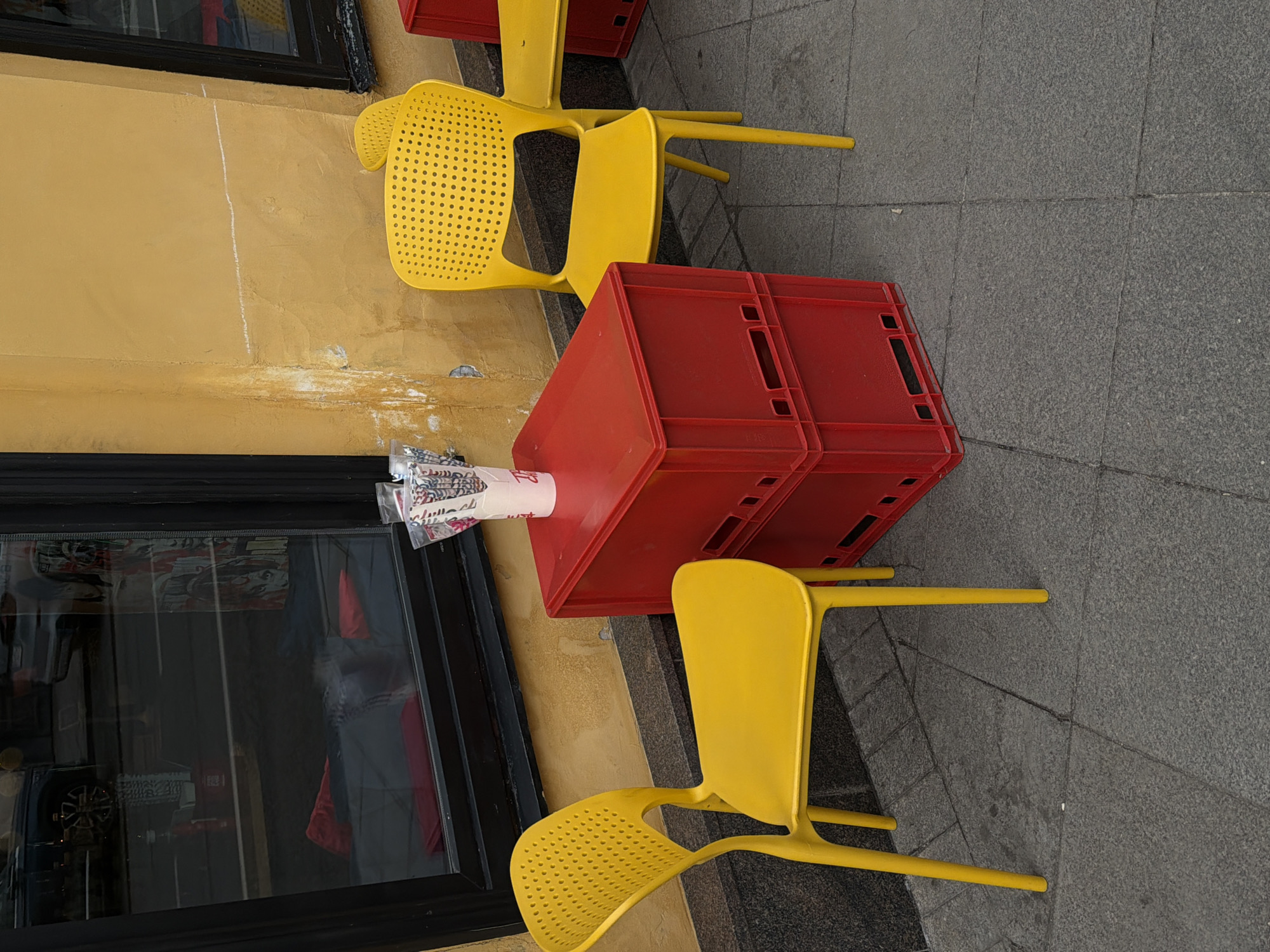 The image presents a scene in what appears to be an outdoor cafe or eatery setting. The location is suggested to be Moscow, Russia but the specific details do not confirm it. The focal point includes two yellow plastic chairs and two red plastic crates stacked atop one another, apparently used as an impromptu table. A disposable paper cup is placed on the red crates. The scene is located beside the wall of a building with a glass window visible. The setting gives an impression of a simple, casual, possibly temporary arrangement. The background suggests an urban environment, with stone pavement beneath the furniture. The scene contains no people and shows no interaction.