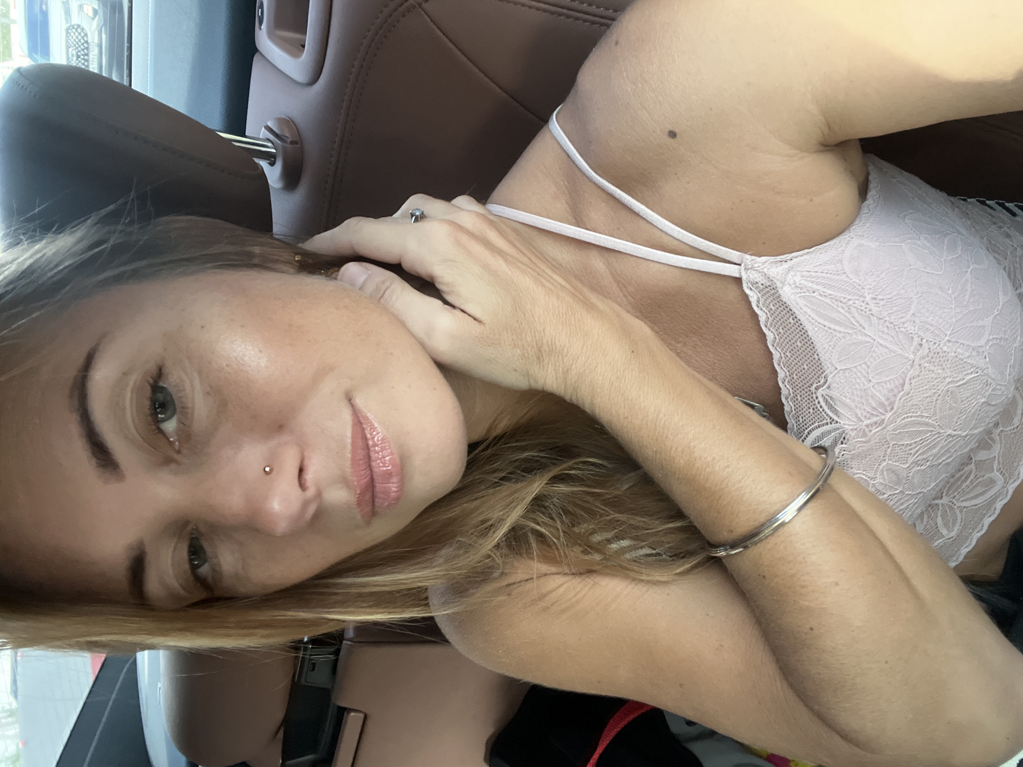 The media file contains a close-up, tilted portrait of a woman in a car. She has blonde hair, green eyes, and is wearing light pink lace bralette with thin straps. Her lips are glossy and a subtle nose ring is visible. She is resting her head on her hand, with her other arm bent and resting on her chest. A silver bracelet is visible on her wrist. The background shows the interior of a car, with brown leather seats and a headrest. The lighting suggests it is daytime. No text is visible in the media file. The woman appears relaxed and at ease.