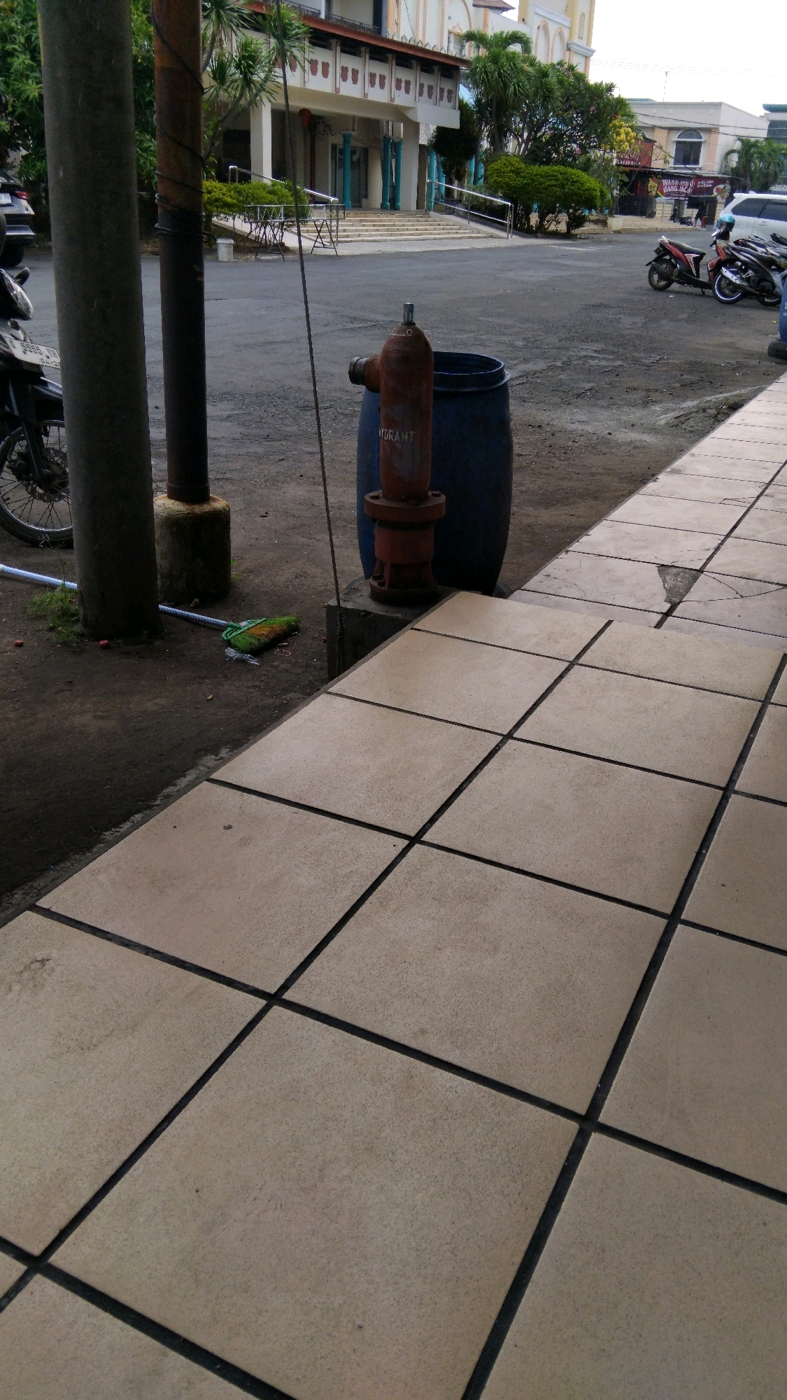 The image depicts an outdoor scene with a paved walkway in the foreground. Behind the walkway, there is a dirt area with two large dark poles, one of which is wrapped with wire. A red fire hydrant stands next to a large blue barrel. Further in the background, a building with white and cream-colored architecture and arched windows is visible. Trees and greenery surround the building. Several motorcycles are parked to the right of the frame, and a white SUV is also present. The sky is overcast, suggesting a cloudy day. The overall environment appears to be an urban setting, possibly a public space or a complex in Pekalongan, Indonesia.