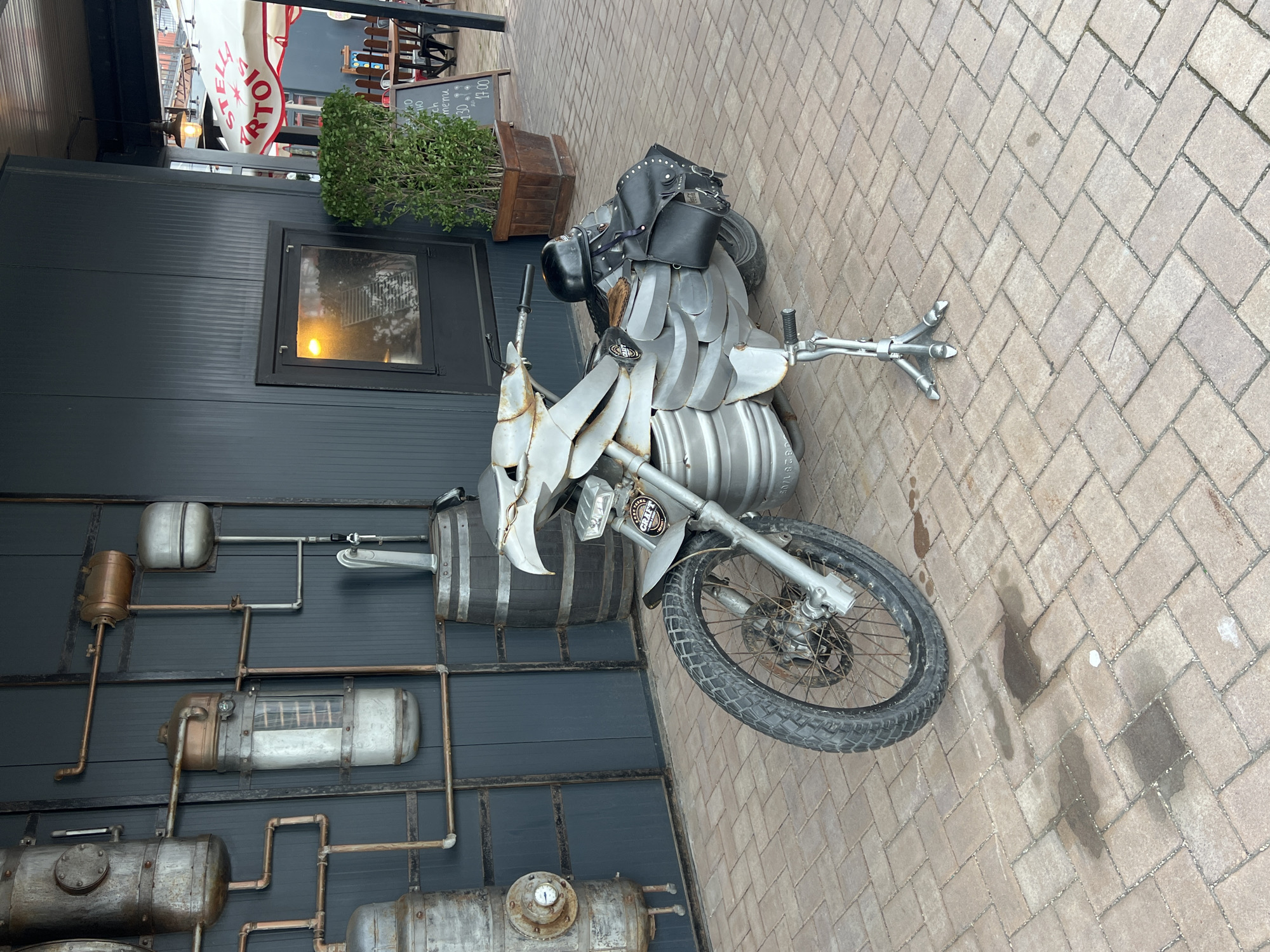 The image captures a custom-built, artistic motorcycle displayed outdoors on a light-colored, herringbone-patterned brick pavement. The motorcycle itself is a prominent subject, constructed from various metal elements, including what appear to be beer kegs or industrial barrels forming its body, and an angular, sculptural front section. It features a black studded saddle and strapped black bags at the rear. The front wheel is spoked, with a disc brake, and the tire has a rugged tread. A unique three-pronged metal kickstand supports the motorcycle. A small badge on the front fender shows partially visible text, including "CRAFT."
Behind the motorcycle is the dark-colored facade of a building. A striking feature of this wall is an elaborate, industrial-themed decoration comprising various interconnected metal pipes, tanks, gauges, and a large wooden barrel, suggesting a steampunk or brewery aesthetic. A rectangular window with reflections is also visible on the wall.
To the right, an outdoor seating area is partially visible under a light-colored awning with "STELLA ARTOIS" printed in red. Wooden tables and chairs are arranged in this area, accompanied by a planter with green foliage and an A-frame chalkboard sign. The text on the sign is largely illegible.
The scene appears to be set during the daytime, with soft, ambient lighting. There are no people present, and no specific activity is taking place; the scene is static. Dark, damp patches are visible on the brick pavement, suggesting spilled liquid or recent moisture. The overall context, including the motorcycle's theme and the building's decor, points towards a pub, bar, or restaurant, consistent with a commercial establishment in a location like Varna, Bulgaria.