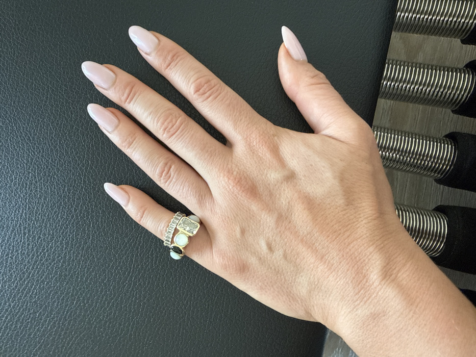 A light-skinned human left hand, exhibiting visible superficial veins on the dorsal surface, is positioned palm-down on a dark, textured surface. The fingernails are manicured with an almond-oval shape and a light pink or nude polish. Two rings adorn the fourth finger: an eternity band composed of multiple small, clear, rectangular stones set in a gold-toned metal, and a larger gold-toned ring featuring a central rectangular clear gemstone flanked by two round, light-colored opaque stones and one small dark stone. The hand and the dark surface are situated above a series of parallel metallic coil springs, which are mounted on dark, cylindrical, padded bases resting on a light-colored floor. These components are consistent with a Pilates reformer or similar fitness equipment. The scene is located in Dubai, United Arab Emirates.