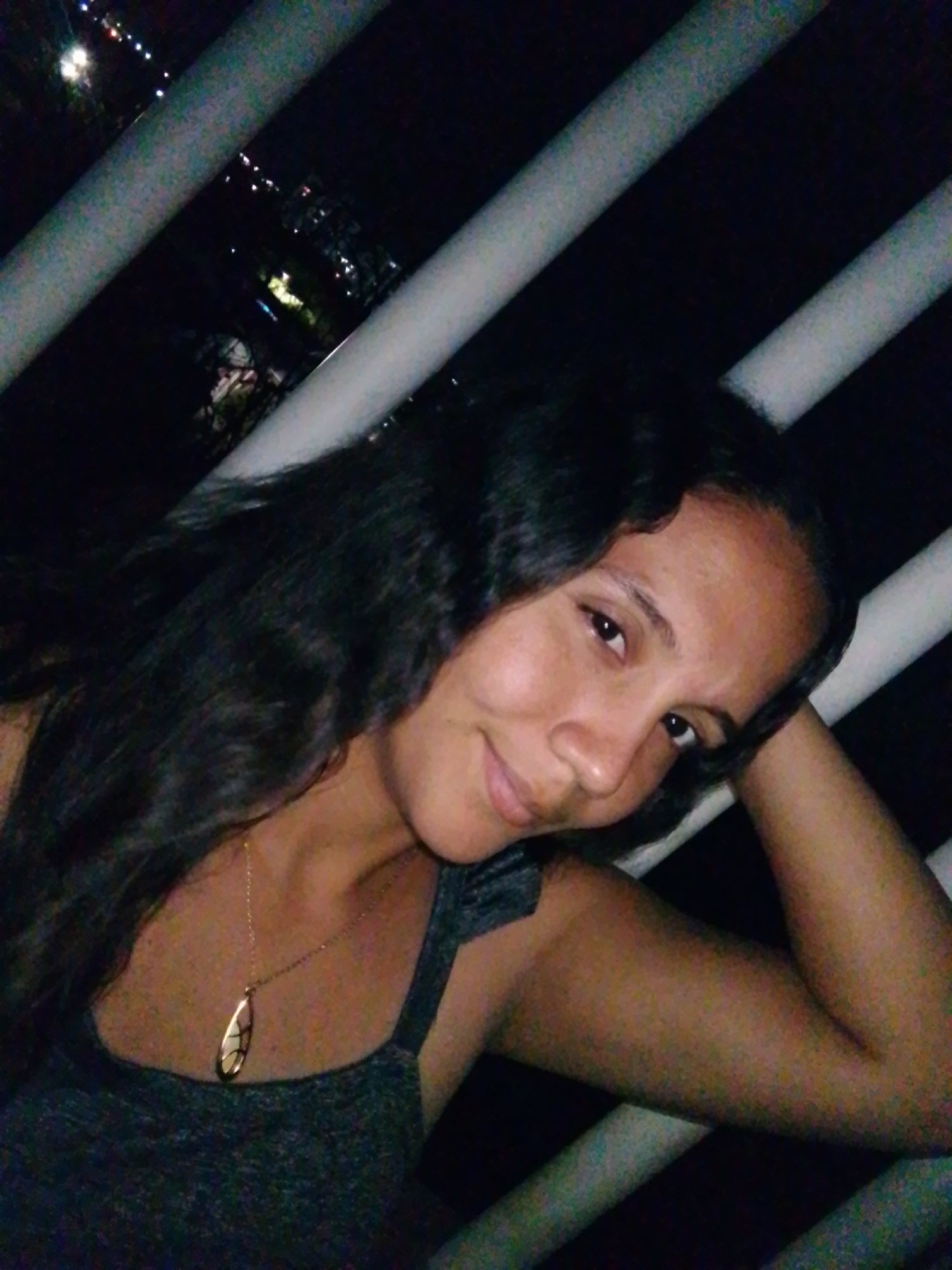 The image displays an adult female photographed at night. She has dark, wavy hair, dark eyes, and a warm skin tone, looking directly at the camera with a faint smile. She is wearing a dark gray tank top and a gold-colored chain necklace with a teardrop-shaped pendant featuring a lighter, possibly iridescent, central design. Her left arm is bent, with her forearm or hand positioned near her head. The background is predominantly dark, traversed diagonally by two light-colored, cylindrical bars or railings. Beyond these railings, blurred artificial lights from a distant populated area are visible against the dark night sky, including scattered streetlights and a prominent bright light source in the upper left. This suggests an elevated viewpoint overlooking a city or town at night. No specific visual identifiers within the image confirm the location as Juan Griego, Venezuela.