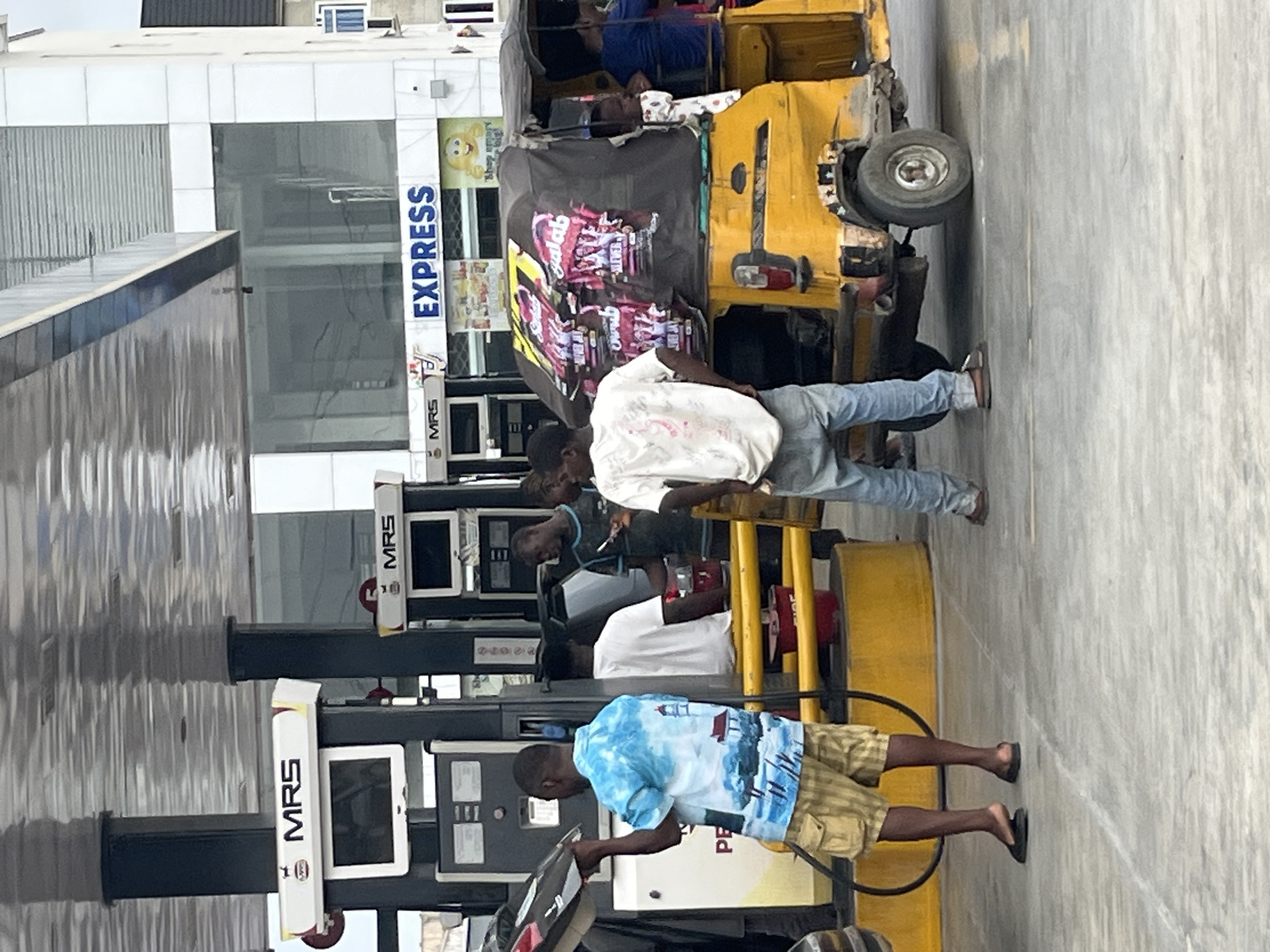 The image depicts a petrol station in Ado, Nigeria. Several people are interacting with the fuel pumps. One man wearing a blue patterned shirt and khaki shorts is fueling a yellow tricycle taxi ("keke"). Two other men are also using the pumps. The station is branded with the "MRS" logo. A building with "EXPRESS" signage is visible in the background.