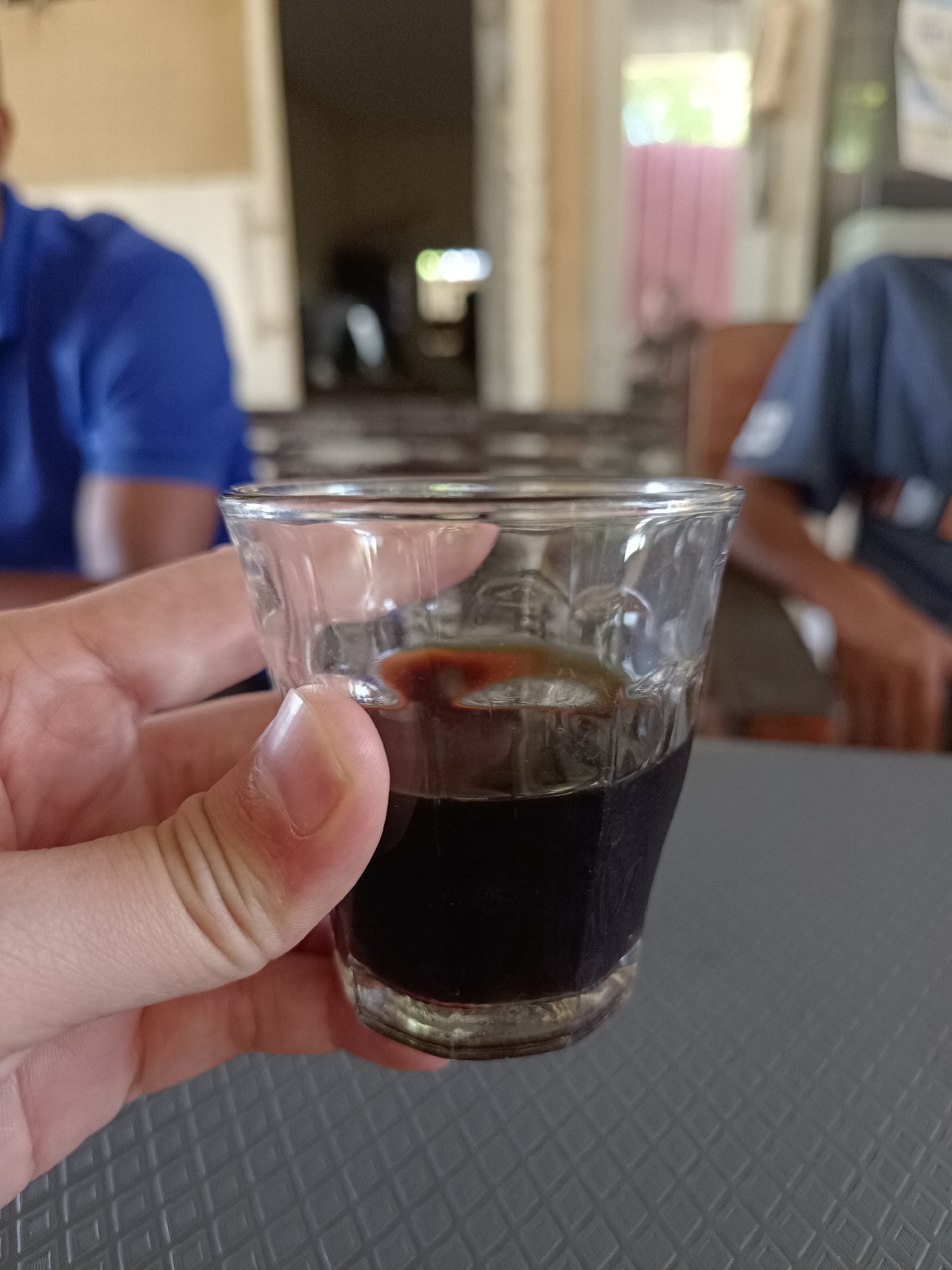 A close-up shot shows a person's hand holding a small glass of dark liquid, possibly coffee or a shot. The glass is clear with a textured pattern near the bottom and contains a dark liquid that reflects a reddish-brown hue near the surface. The hand is positioned on the right side of the glass, with fingers gently gripping the sides. In the blurred background, two individuals can be seen wearing blue shirts. The environment appears to be indoors, possibly a cafe or dining area, with natural light filtering in from an open doorway. The surface below the glass is a textured gray tabletop.