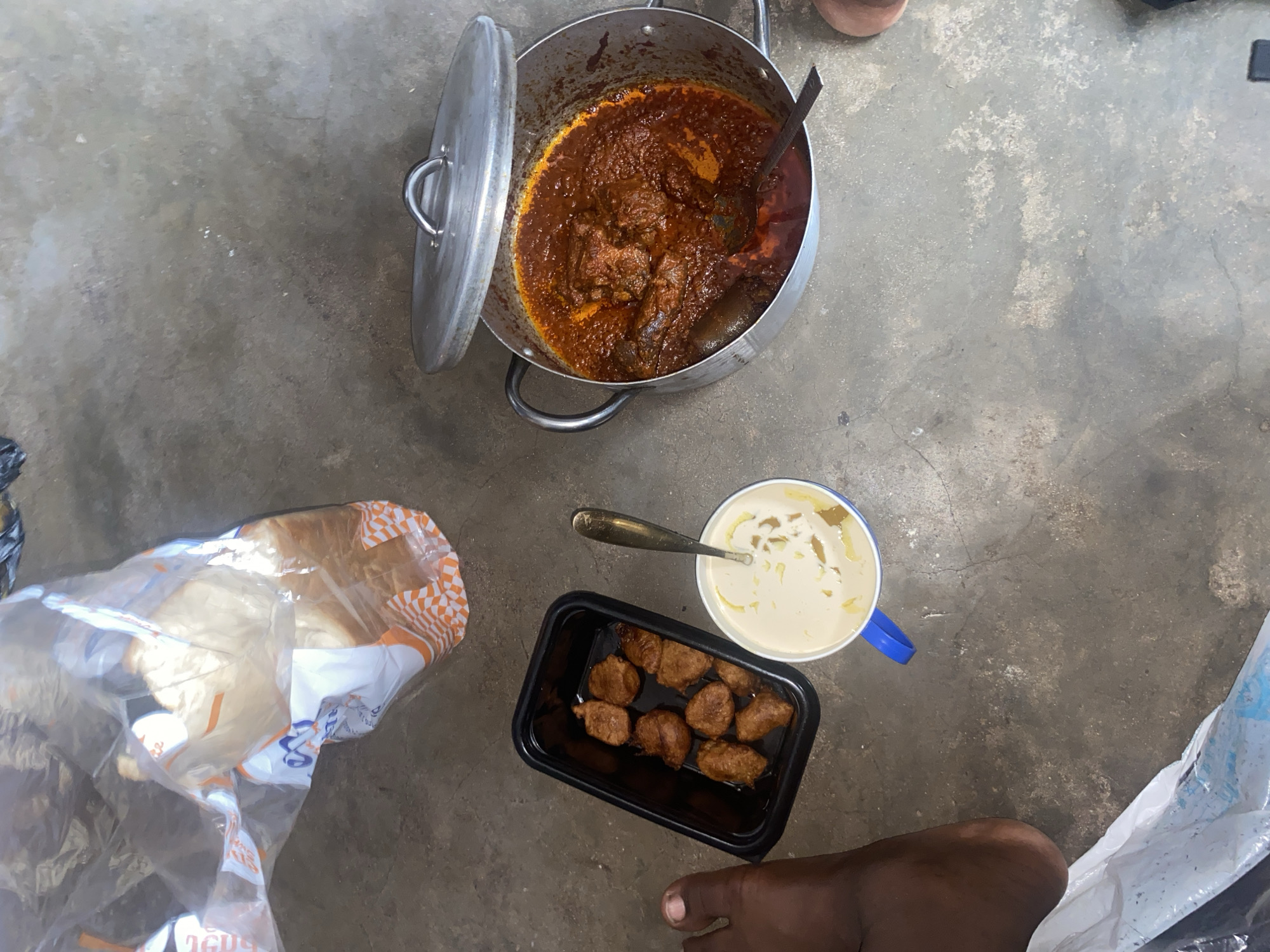 On a concrete surface in Lagos, Nigeria, a partially opened stainless steel pot holds a dark red stew containing meat. A silver spoon rests in a white ceramic mug with a blue handle, containing a light-colored substance. A black rectangular container holds small, round, brown food items. A loaf of bread is partially enclosed in a plastic bag with orange and white patterned trim. Human feet are visible on the right side of the image.