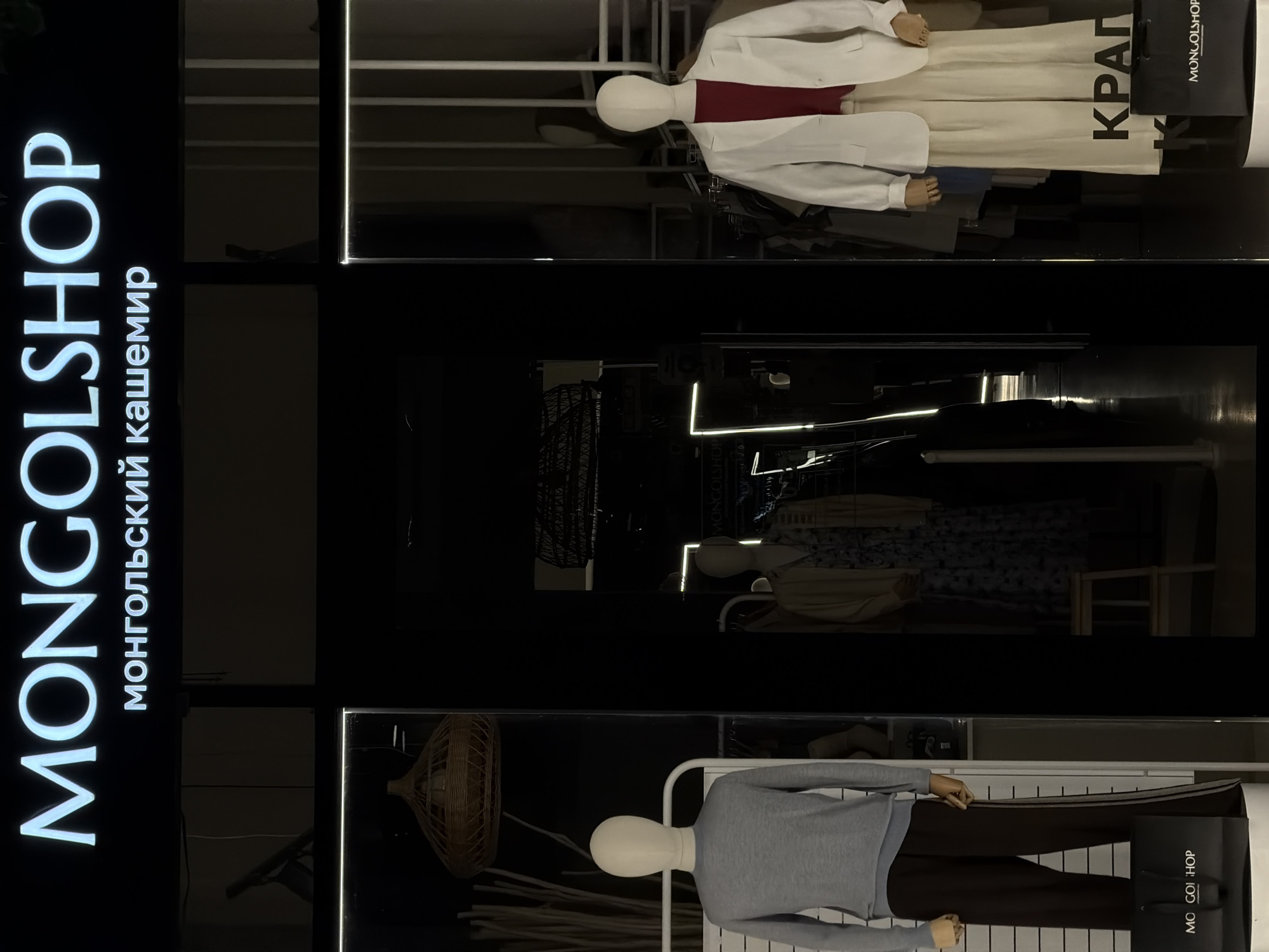 The image shows a storefront in Moscow, Russia, featuring the "MONGOLSHOP" sign, specializing in Mongolian cashmere. The storefront has several display windows, each showcasing mannequins dressed in clothing. The top window features a mannequin wearing a white shirt and pants with the word "KRAP" printed on the pants, along with a shopping bag also bearing the "MONGOLSHOP" logo. The middle window displays mannequins in darker, less distinct clothing, while the bottom window shows a mannequin in a grey sweater and dark pants, again with a "MONGOLSHOP" shopping bag. A decorative woven object hangs in the corner of the bottom window. The scene is taken at night, emphasizing the illuminated signage and displays.