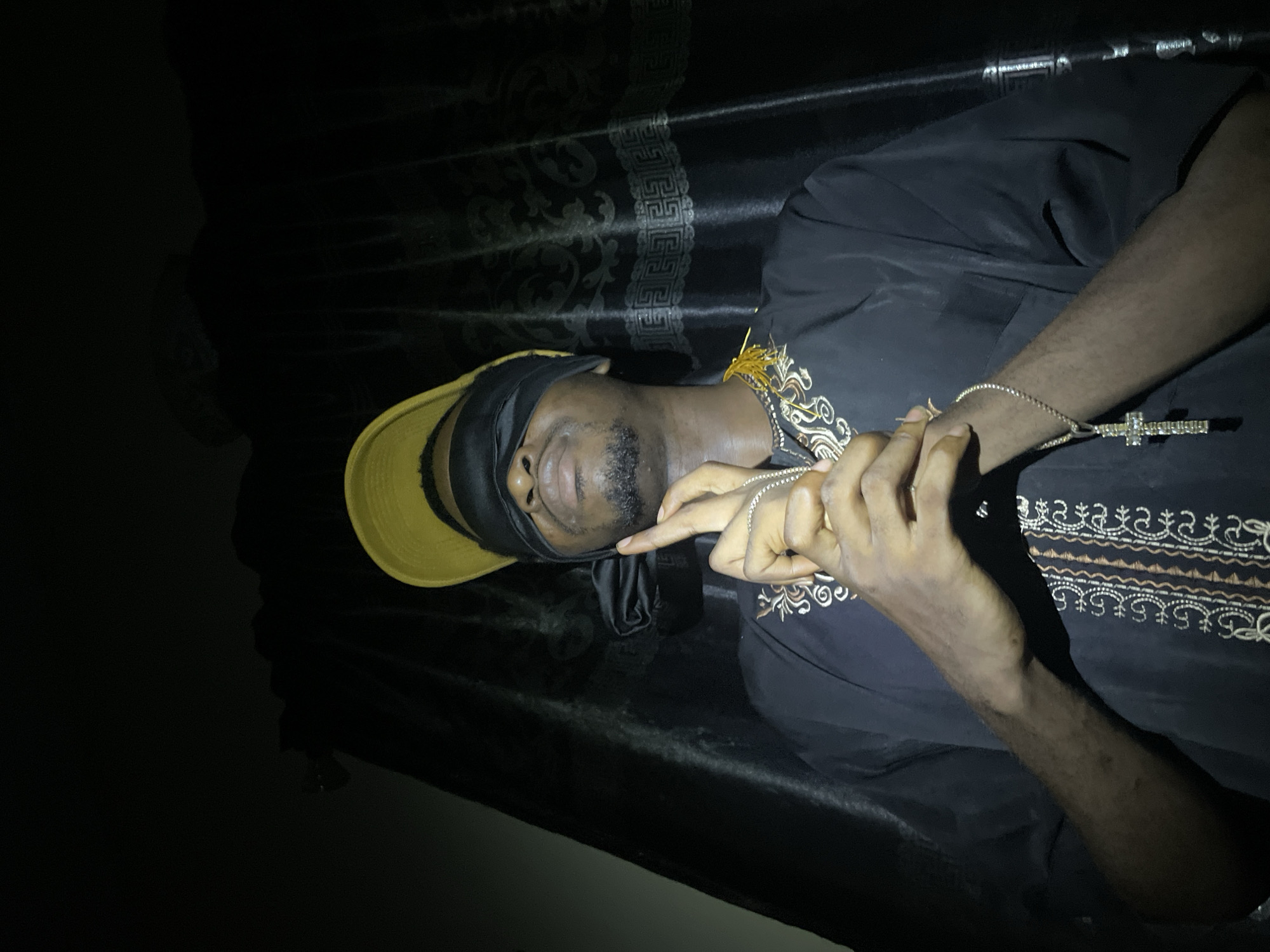 A dark-skinned man is lying down on what appears to be a black surface with a patterned backdrop, possibly fabric. He is wearing a gold-colored baseball cap with a black band covering his eyes. He is wearing a black shirt with gold embroidery along the collarbone and sleeve. He has a gold bracelet on his left wrist and is holding two silver necklaces in his hands, one with a large cross pendant. The setting is dimly lit. The location, Lokoja, Nigeria, is mentioned solely in the prompt and is not discernible from the image itself.