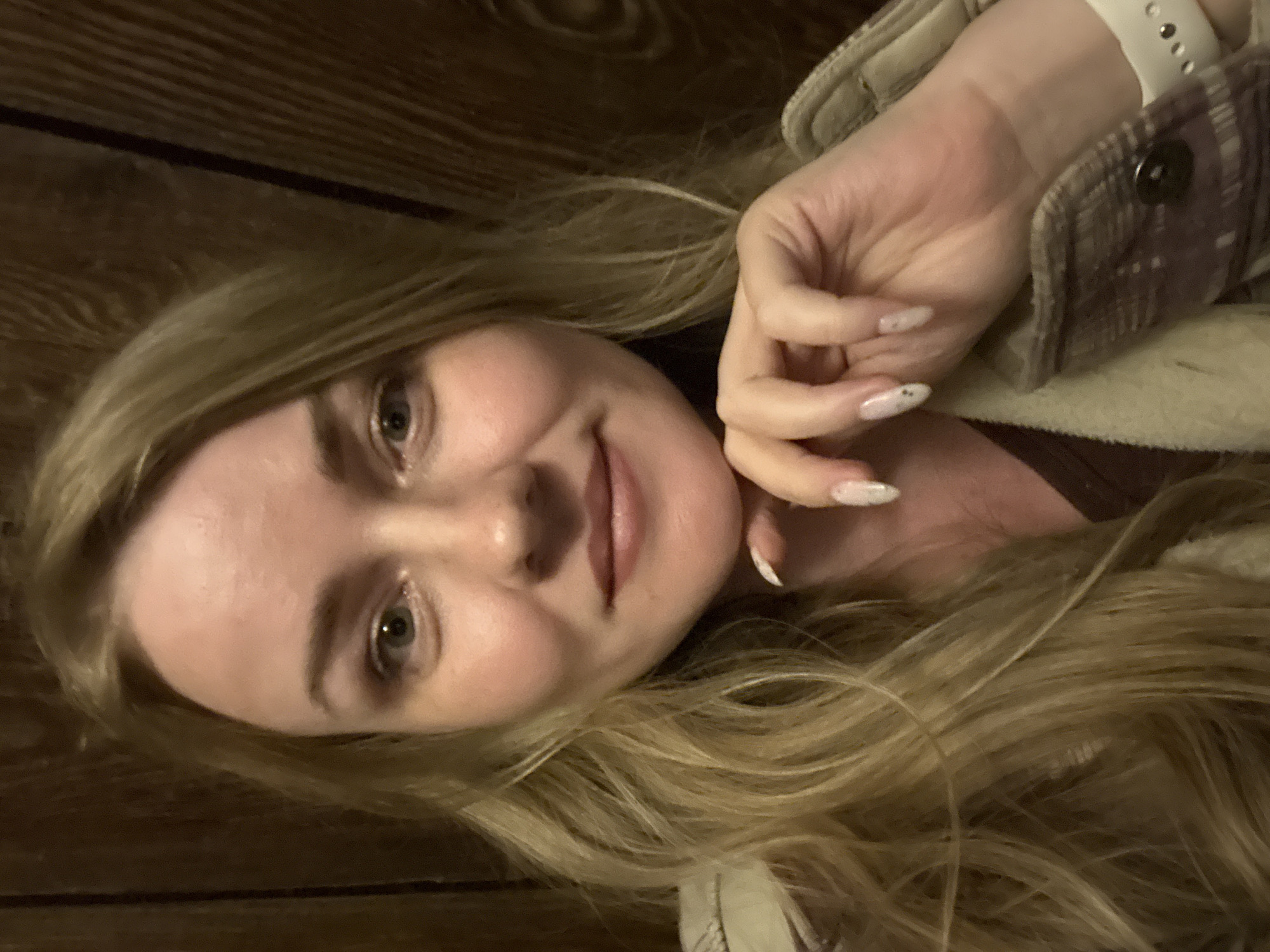 A portrait of a young woman with long blonde hair, fair skin, and light eyes is angled against a dark, possibly wooden, wall. She is smiling slightly and has her hand raised near her face, displaying a manicure with light-colored polish. She is wearing a beige jacket and a smartwatch with a white band on her wrist.
It is impossible to confirm the location to be the city of Chaikovsky, Russia.