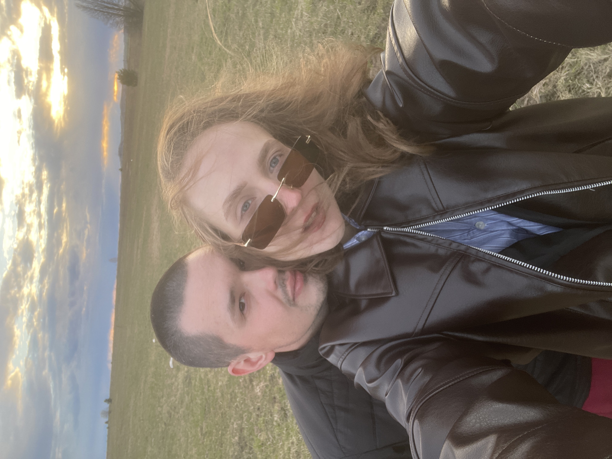 This image captures a couple, a man and a woman, posing closely for a photo outdoors in what appears to be a vast open field in Kueda, Russia, during either sunrise or sunset. The scene is illuminated by the "golden hour," with warm, golden light bursting through clouds on one side of the sky.
The woman, positioned slightly in front, has long, wind-blown, light brown hair. She is wearing stylish rectangular sunglasses with brown-tinted lenses, revealing glimpses of her light blue eyes. She sports a subtle smile and is dressed in a black leather jacket over a blue and white striped shirt.
The man is situated slightly behind and below her. He has short, dark hair and light stubble on his face, displaying a neutral to gently smiling expression. He is also wearing a black leather jacket.
The background showcases an expansive green field extending to a distant horizon dotted with sparse trees and possibly hills. The sky, a prominent feature, is filled with scattered clouds beautifully lit by the low sun. The overall atmosphere conveys a sense of affection and tranquility, capturing a shared moment between the couple in a picturesque natural setting. There is no visible text in the image.
