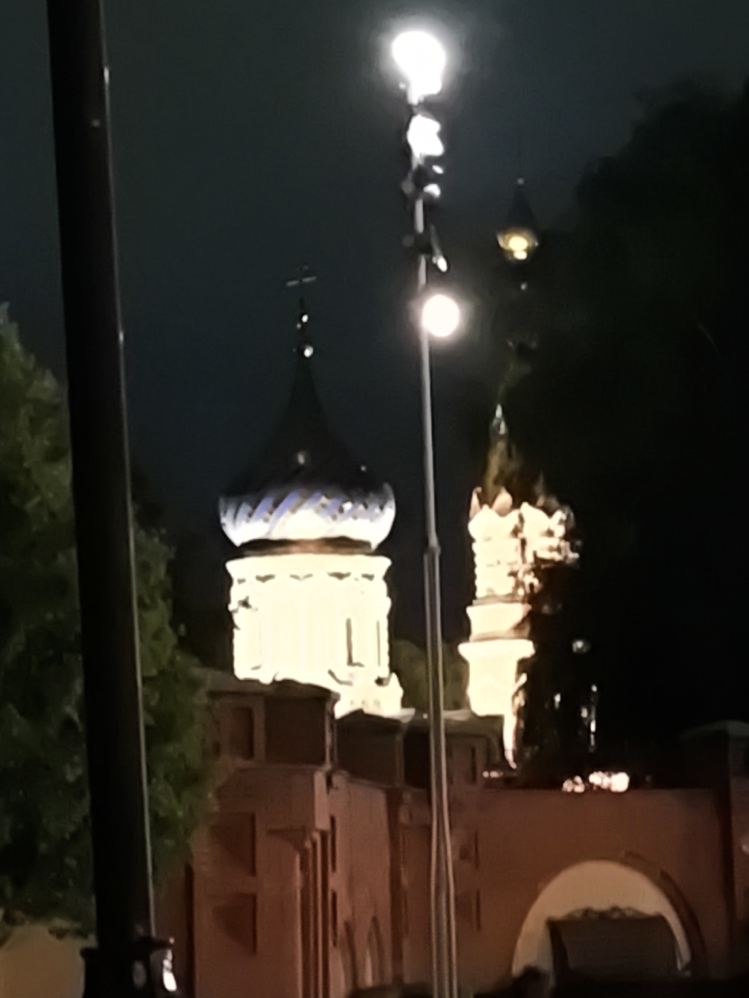 This nighttime image depicts St. Michael's Cathedral (Mikhailovsky Cathedral) in Izhevsk, Russia, viewed from a lower perspective. Dominating the center-left is the brightly illuminated white-stone structure of the cathedral, topped by a distinctive blue onion dome patterned with white, surmounted by an Orthodox cross. To the immediate right of the cathedral, a similarly illuminated structure with decorative elements is partially visible, likely an adjacent part of the cathedral complex. In the foreground, a tall, dark streetlamp pole with two illuminated spherical lights, one at the top and one mid-way down, stands prominently in the center, casting a bright glow. Another dark pole is partially visible on the far left. The lower section of the image reveals a red-brown building or wall structure with arched openings and rectangular features, also illuminated by warm light. Dark foliage is visible on the left and right sides of the frame, silhouetted against the illuminated structures and the dark night sky. No people are visible.
