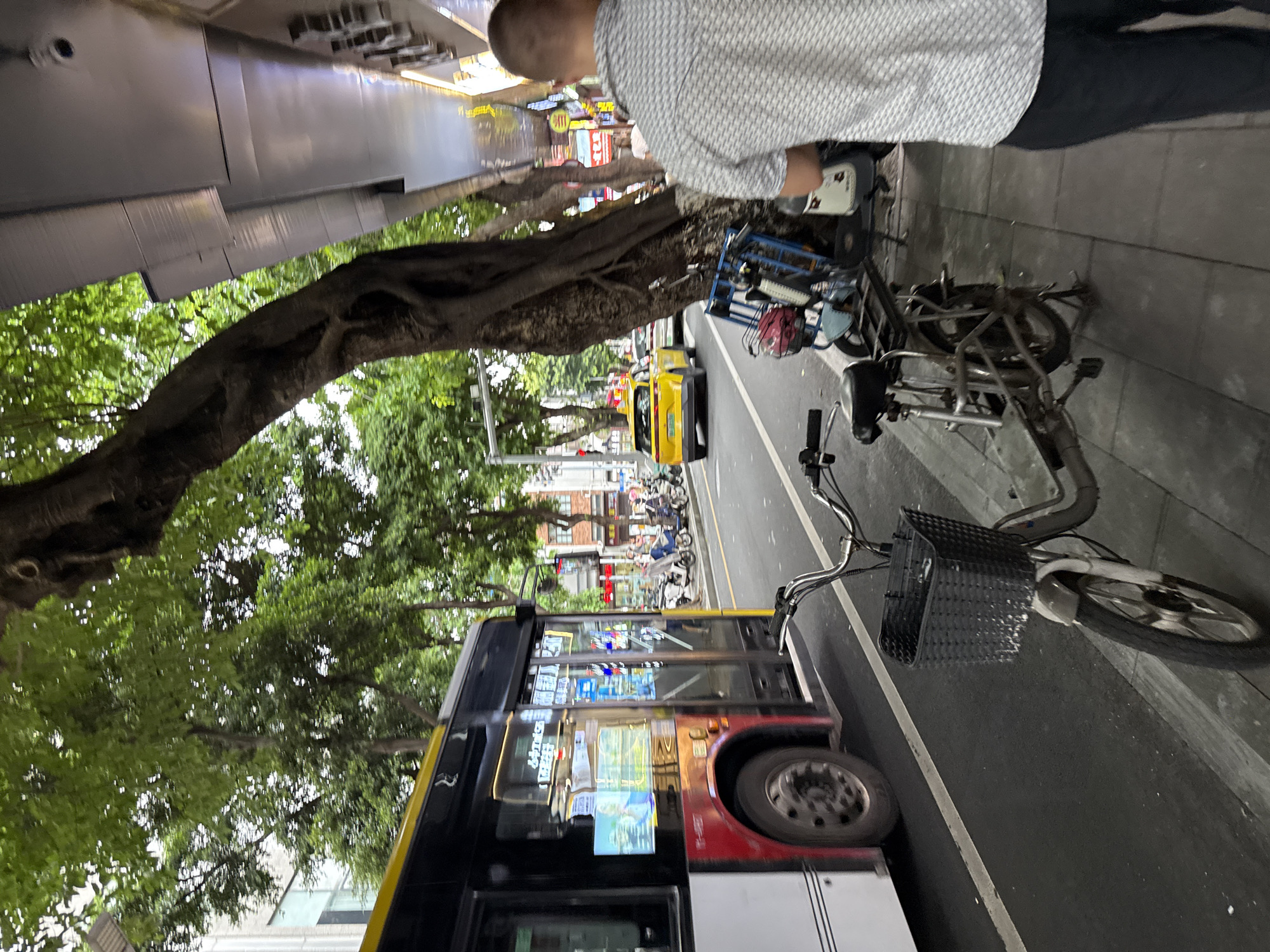 A man in a patterned shirt walks on a sidewalk next to a street in Liwan District, China. A yellow taxi drives by on the road, followed by a bus. Several electric scooters are parked on the sidewalk. The street is lined with trees and buildings.