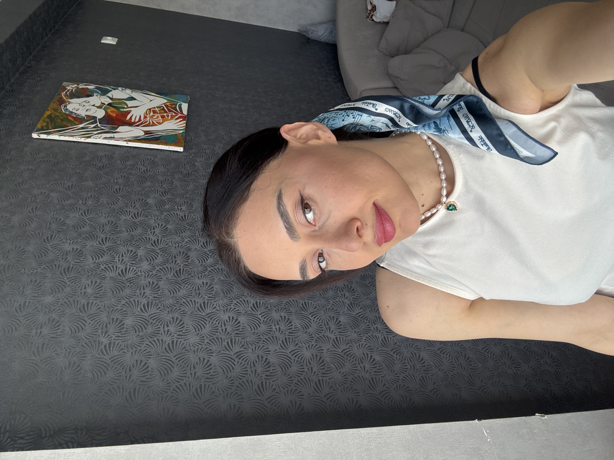 A young woman poses for a vertical selfie indoors in Tbilisi, Georgia. She has dark hair pulled back and is wearing a white short-sleeved top. Around her neck, she wears a white pearl necklace with a small green, heart-shaped pendant, and a blue and white patterned silk scarf. Visible text on the scarf appears to include "Bluetbl" and "Street."
The woman has defined eyebrows, brown eyes, and pink-red lipstick. She looks directly at the camera with a gentle expression.
The background features a dark gray wall or surface with a subtle, repeating embossed pattern resembling stylized fans or shells. To her left, a rectangular painting rests on this surface, depicting two abstract figures, possibly women, outlined in white against a vibrant background of red, yellow, green, and blue. A light gray sofa or armchair is visible in the upper right background, suggesting a home or studio setting. The scene is brightly lit, likely by daylight.