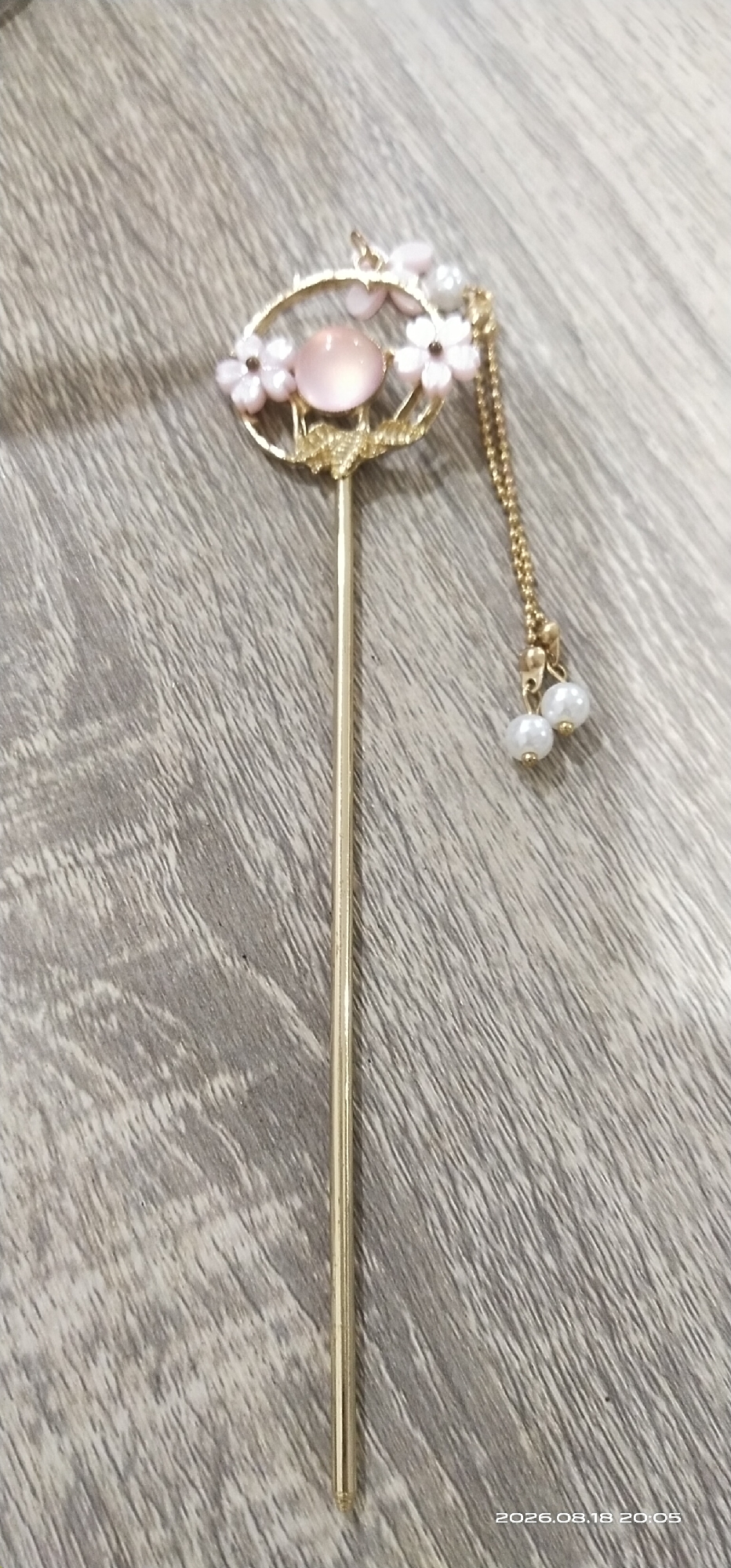 A close-up, slightly angled shot shows a gold hairpin with a circular decorative top adorned with small pink flowers, a light pink gemstone, and a gold chain with two white pearl-like beads. The hairpin is placed on a wooden surface with a visible wood grain texture. The timestamp "2026.08.18 20:05" is visible in the lower right corner of the image.