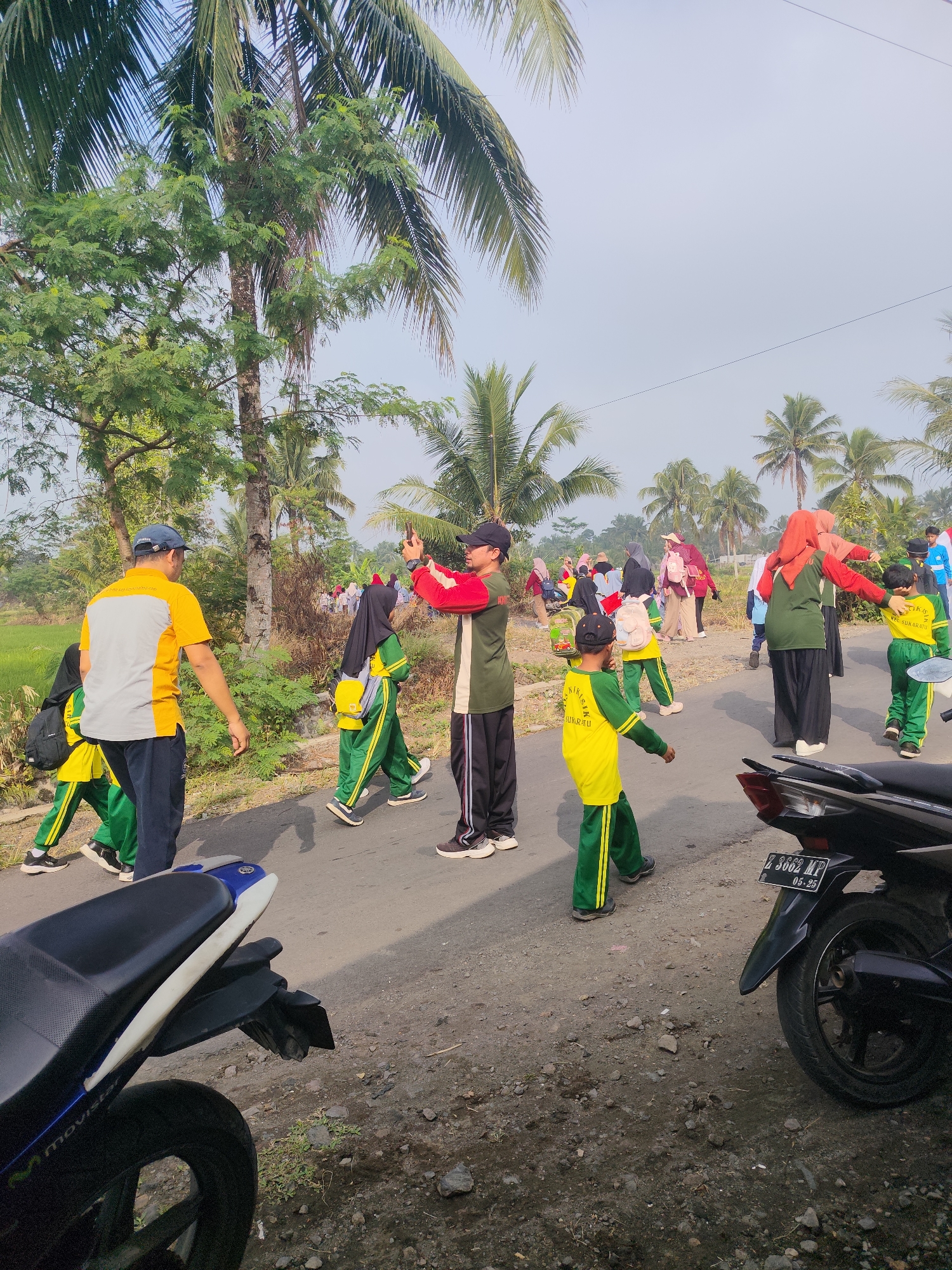In Sukaratu, Indonesia, a group of people, including children in matching green and yellow uniforms, are walking along a paved road. The children appear to be part of a school group, as indicated by their attire and backpacks. Some adults are present, accompanying the children and observing the activity. One adult is taking a photo with his phone, while another is engaged in conversation with a child. The environment is rural, with palm trees lining the road and lush vegetation visible in the background. A motorcycle is parked on the side of the road, adding to the everyday scene. The sky is clear and the overall atmosphere suggests a community event or an organized outing for the children.
