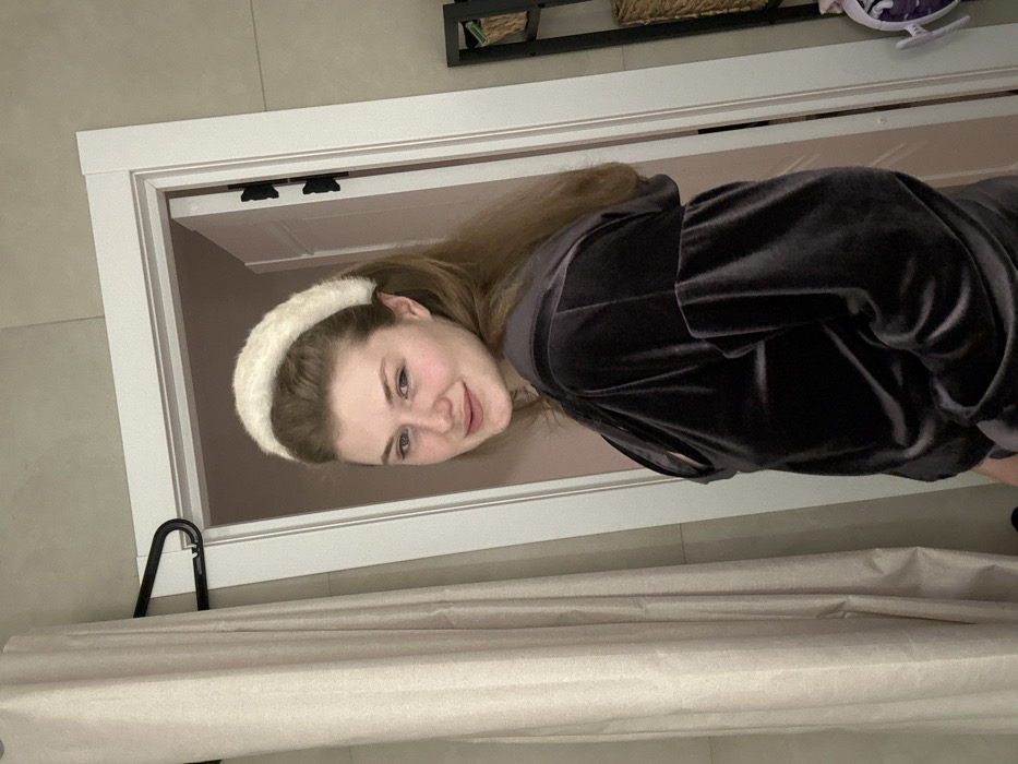 A young woman with fair skin and light brown hair is wearing a fluffy, white headband and a dark gray velvet hooded jacket. She is positioned within a doorway, with a beige shower curtain in the foreground and a glimpse of a room behind her. The photo is oriented sideways.