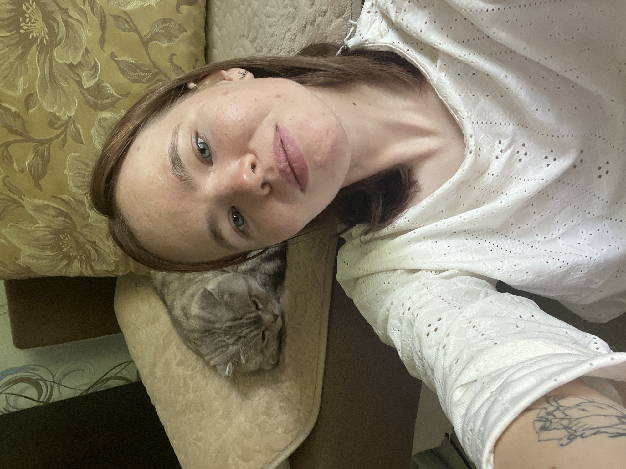 The image captures a young woman and a domestic cat in a relaxed indoor setting, likely in Chaikovsky, Russia. The woman, with brown hair and blue eyes, takes a selfie from a high angle, looking directly at the camera with a neutral, slightly smiling expression. She has light makeup, including pink lipstick, and a small silver ear piercing. A black-outlined tattoo featuring an abstract or stylized design is visible on her right forearm. She is wearing a white top with intricate eyelet or perforated detailing.
The woman is reclining on a couch, partially visible with a patterned cushion featuring a gold and brown floral design, and a lighter beige, textured cushion next to her. A light gray tabby cat is curled up and appears to be sleeping peacefully on the beige cushion, near the woman's head. In the background, a wall with light-colored, swirly patterned wallpaper is faintly visible. The scene suggests a calm, domestic atmosphere. There is no visible text in the image.