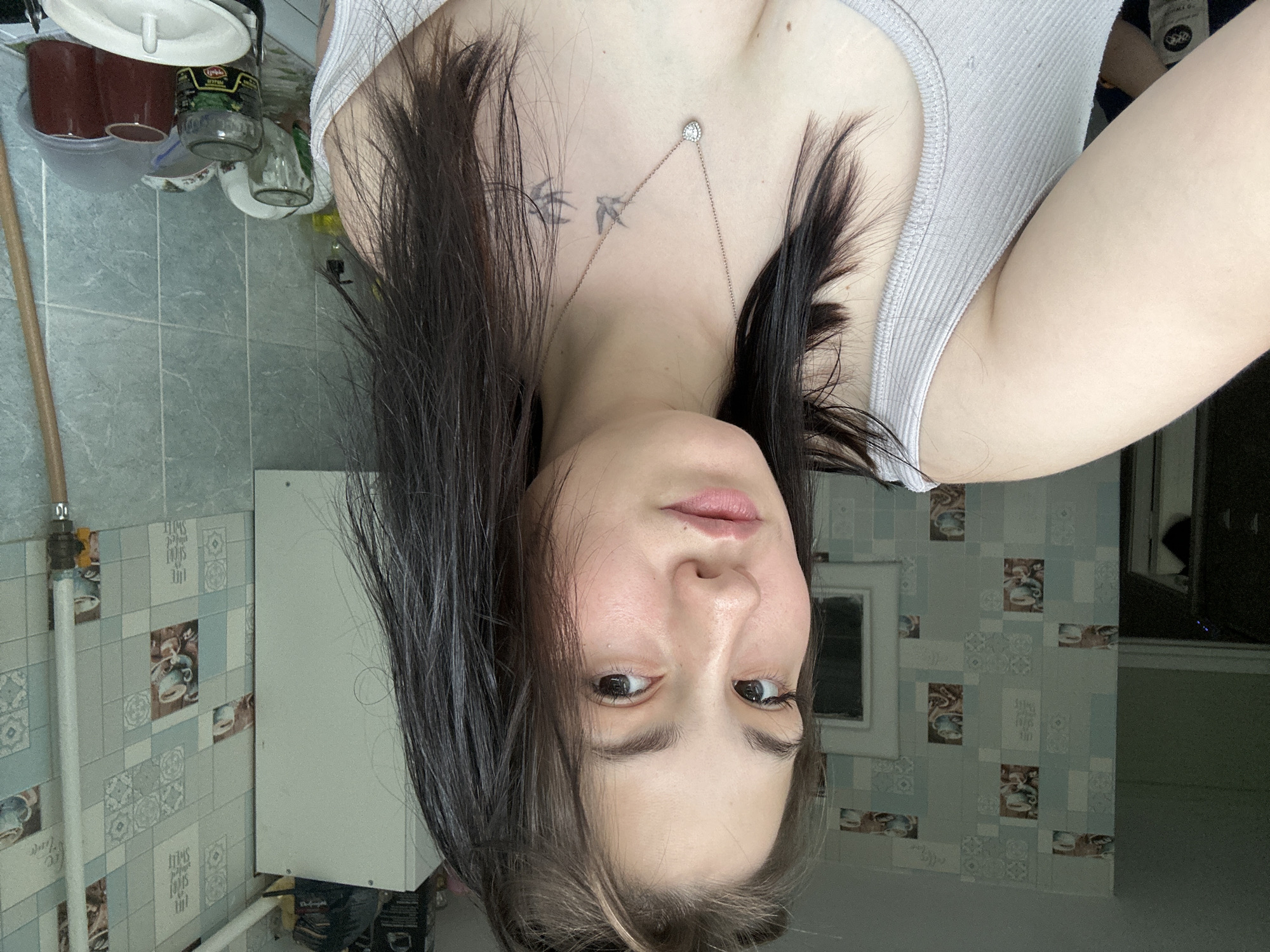 The media file features a young woman in a close-up, upside-down shot. She has dark hair that falls around her face and shoulders, and she is wearing a light-colored, ribbed tank top. A delicate necklace with a circular pendant rests on her collarbone, above a tattoo of two birds in flight. Her face is turned slightly, with her eyes looking towards the viewer, and her lips are slightly parted.
The background appears to be a bathroom or kitchen area, with light blue and white tiled walls featuring a repeating pattern of small images, possibly of teacups or coffee cups. A white cabinet or appliance is visible on the left side of the frame, and a plumbing fixture is attached to the wall. On a shelf above, there are several containers, including a glass jar with a label that seems to read "Lyubimaya" in Cyrillic script, which translates to "Beloved" in Russian. Another jar and some dark red cups are also visible.
The lighting suggests it might be daytime, though no specific weather cues are apparent. The overall impression is an informal, personal snapshot, possibly taken in a domestic setting in Chaikovsky, Russia. There are no other people or animals visible in the frame, and the focus is entirely on the woman and her immediate surroundings. The image is rotated 180 degrees, making it appear upside down.