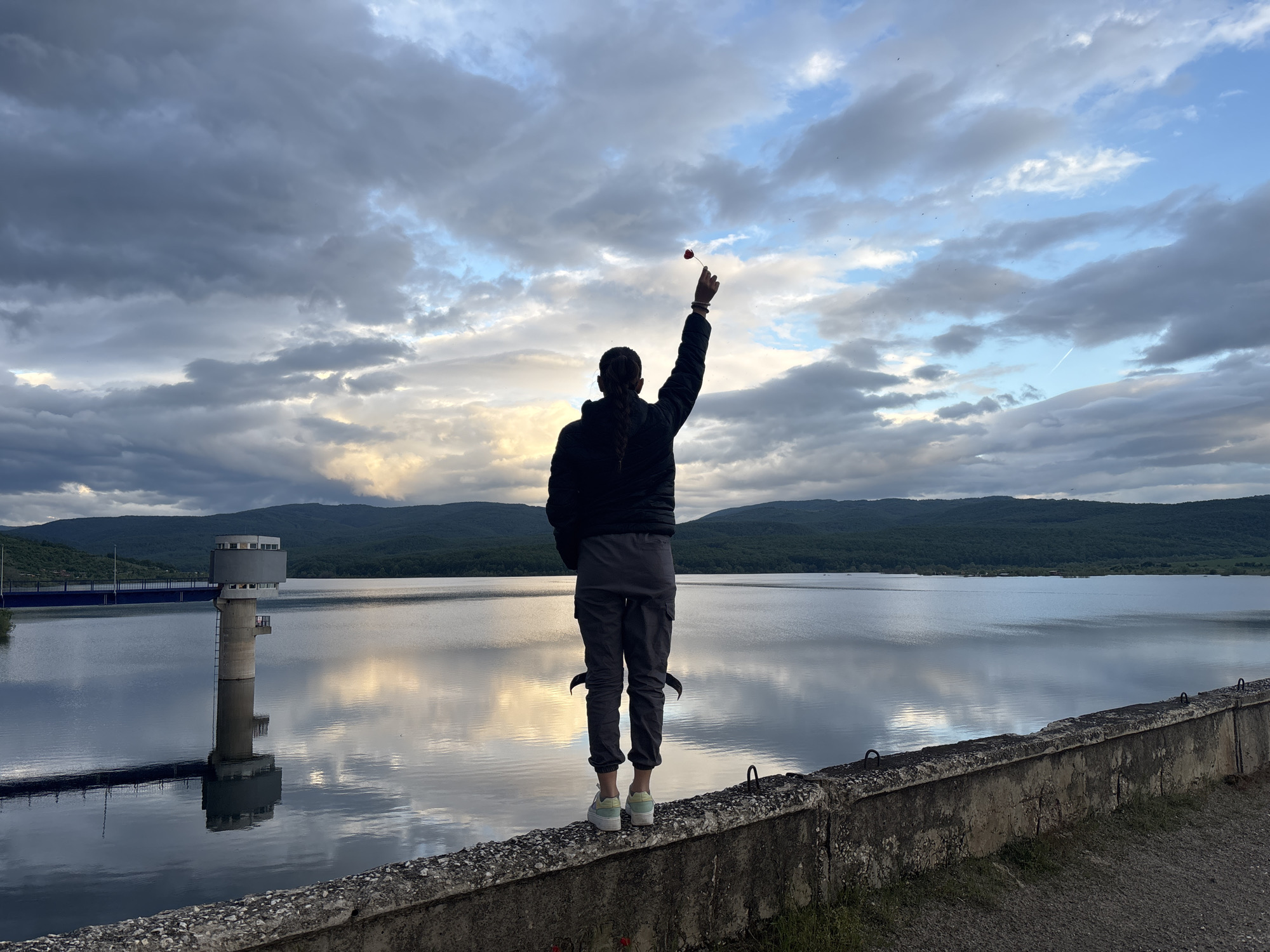A person stands on a concrete ledge, raising an arm with a small red object in their hand against a backdrop of a large body of water and overcast sky. The person is seen from the back, wearing a black jacket, gray pants, and light-colored sneakers. Their hair is in a braid. The body of water reflects the sky and surrounding landscape. To the left is a tall, cylindrical tower with windows. In the background are rolling hills covered in vegetation. The location is likely the Ognyanovo reservoir in Bulgaria, recognizable by the tower structure. The person is standing on a low concrete wall next to the water.