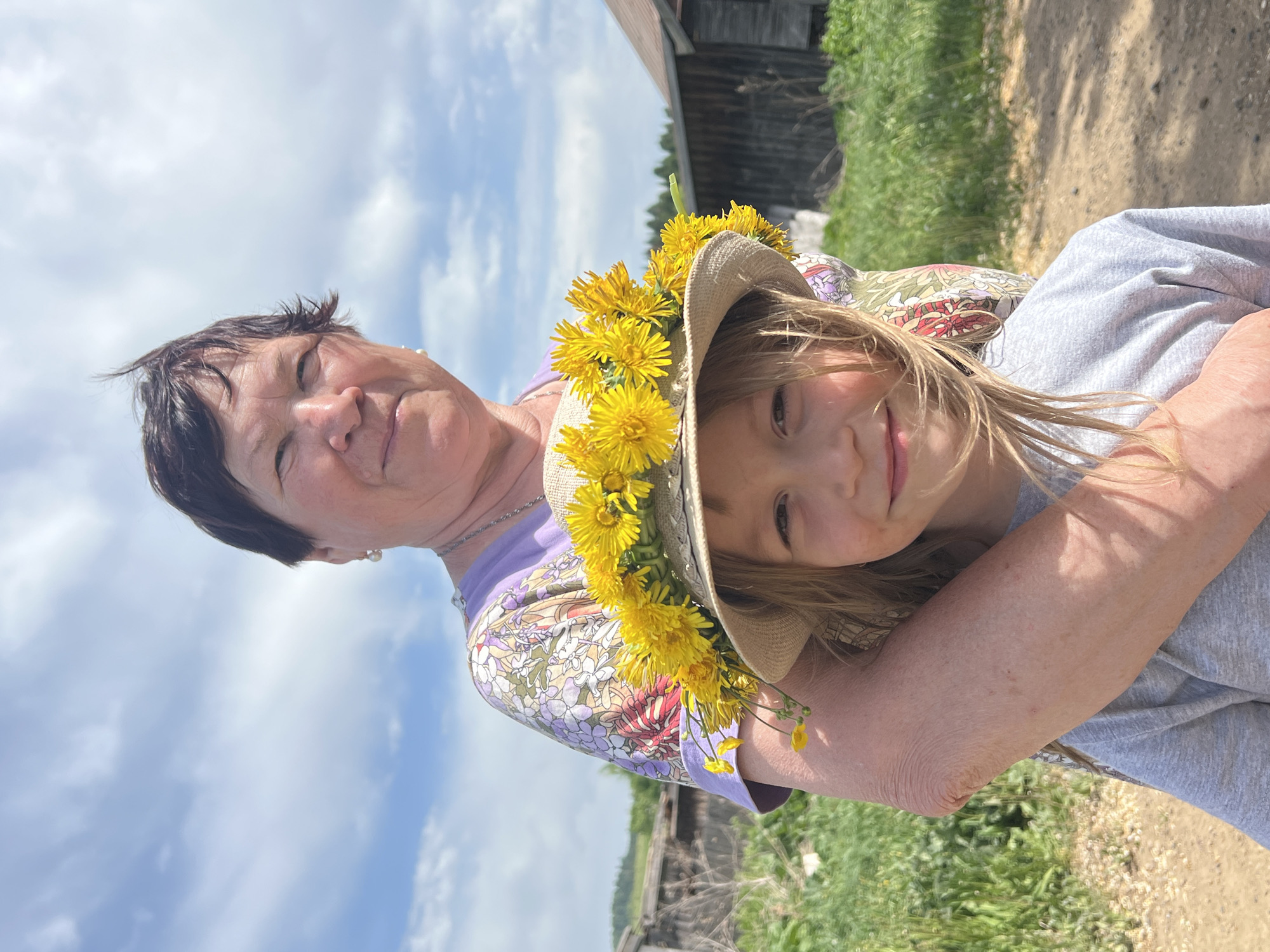A woman is holding a child wearing a hat decorated with yellow flowers. The woman is wearing a floral shirt. The background includes a partly cloudy sky, green vegetation, a dirt path, and a dilapidated wooden structure. The location is likely rural. The setting may be in Panteleevka, Russia.