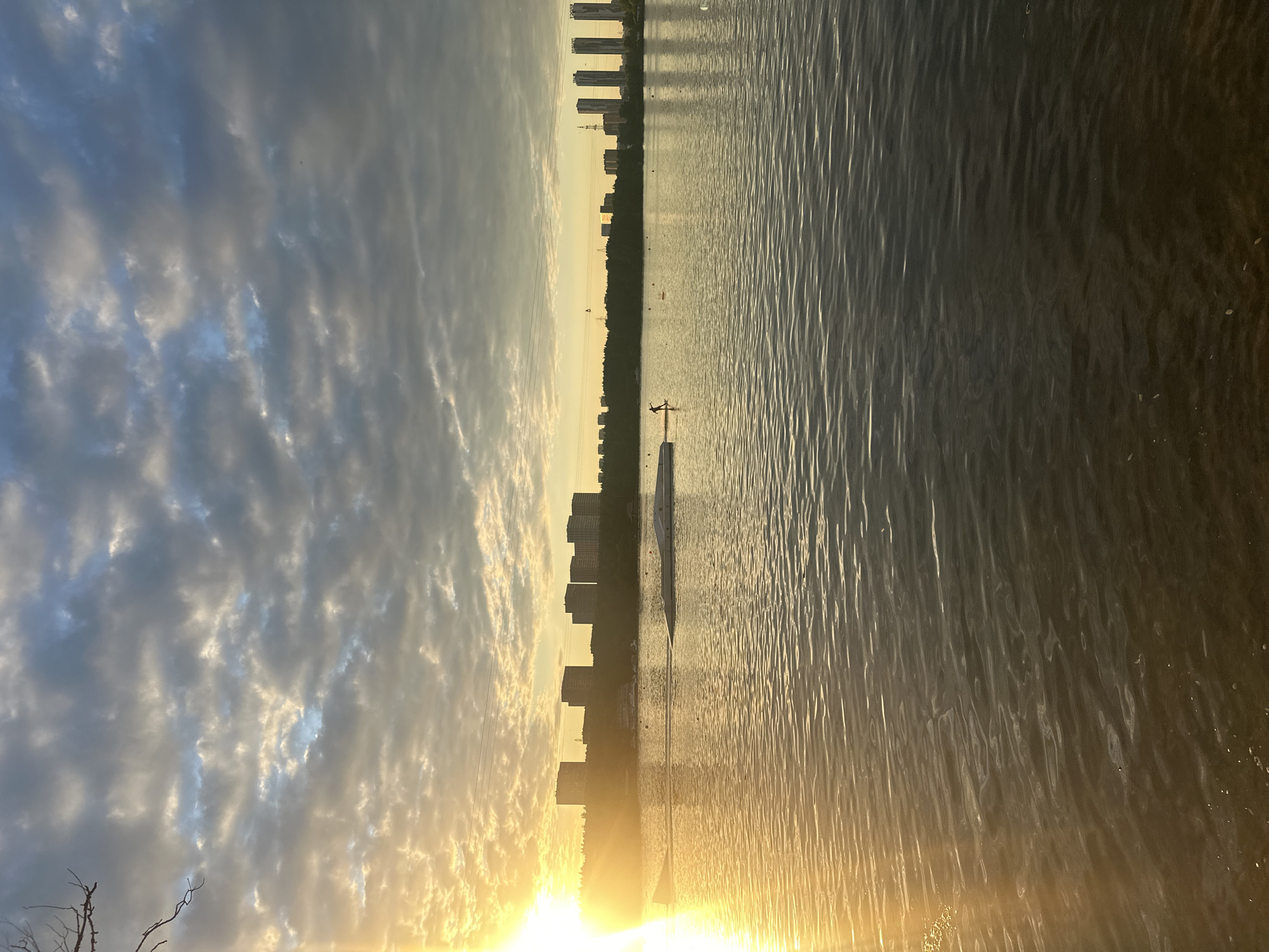 A sunset over a wide body of water with a cityscape in the distance. The sun, a bright golden orb, is low on the horizon, casting a warm glow across the scene. The sky is filled with a mix of dramatic, textured clouds, ranging from deep blues and grays to bright whites and yellows where the sunlight catches them.
The water's surface is rippled, creating a shimmering texture that reflects the sky and the warm light. A dark line of trees forms a barrier between the water and the city. The cityscape is composed of several tall, modern buildings that rise above the trees, silhouetted against the bright sky. Some of these buildings appear to be apartment complexes or office towers.
In the foreground, on the water, there is a slender white structure, possibly a pier or a platform, with a dark figure standing on it. The figure is silhouetted and appears to be engaged in an activity, possibly sports or recreation, given the context of the water and the distant city. There are also some faint reflections of lights or structures on the water in the far distance, near the cityscape. The overall mood is serene and contemplative, with the beauty of nature and urban development coexisting. The time of day is clearly sunset, and the weather appears to be clear with some cloud cover. The location cues of tall buildings and a prominent body of water suggest an urban environment, possibly a river or a large lake within Moscow, Russia, as suggested by the location context. No visible text is present in the image.