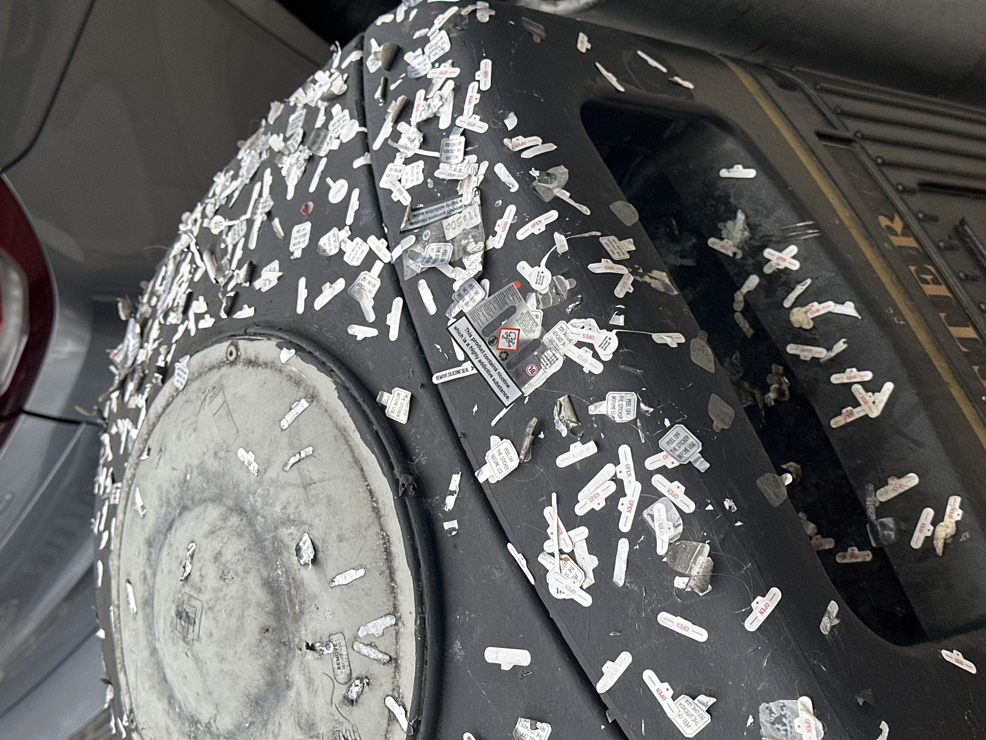 The image depicts a heavily vandalized black object, possibly a piece of street furniture, densely covered with small white stickers that say 'OPEN' and 'PEEL OFF THE STICKER BEFORE USE'. Some stickers are partially torn or peeling. There's a larger sticker with warning symbols, including a skull and crossbones, and text indicating the product contains nicotine and is highly addictive. The object has a textured surface and appears to be part of a larger structure, potentially a bin or a box. The background shows a blurred silver vehicle, suggesting the location is likely a street or parking area. Without additional information, pinpointing the location in London is impossible.