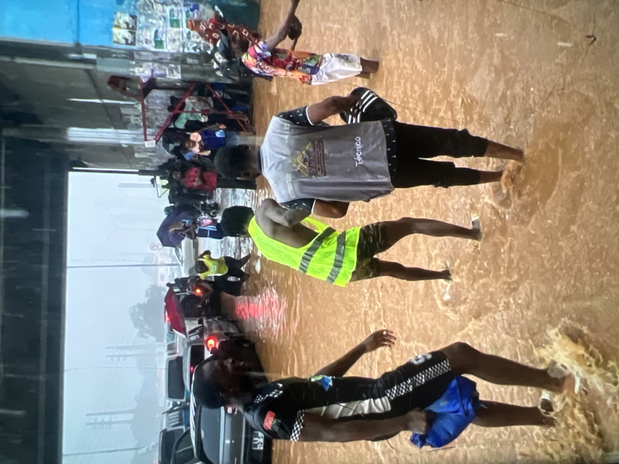 The image depicts a flooded urban street in Lagos, Nigeria, during a heavy downpour. Cars are submerged in murky brown water, creating traffic congestion. Several individuals are wading through the floodwaters, some appearing to be assisting others. One person in the foreground, wearing a black and white checkered shirt and shorts, carries a blue bag. Another person in a bright yellow vest is also visible. In the background, people are using umbrellas and some vehicles are partially submerged. The scene suggests a significant disruption to normal city life due to the severe flooding.