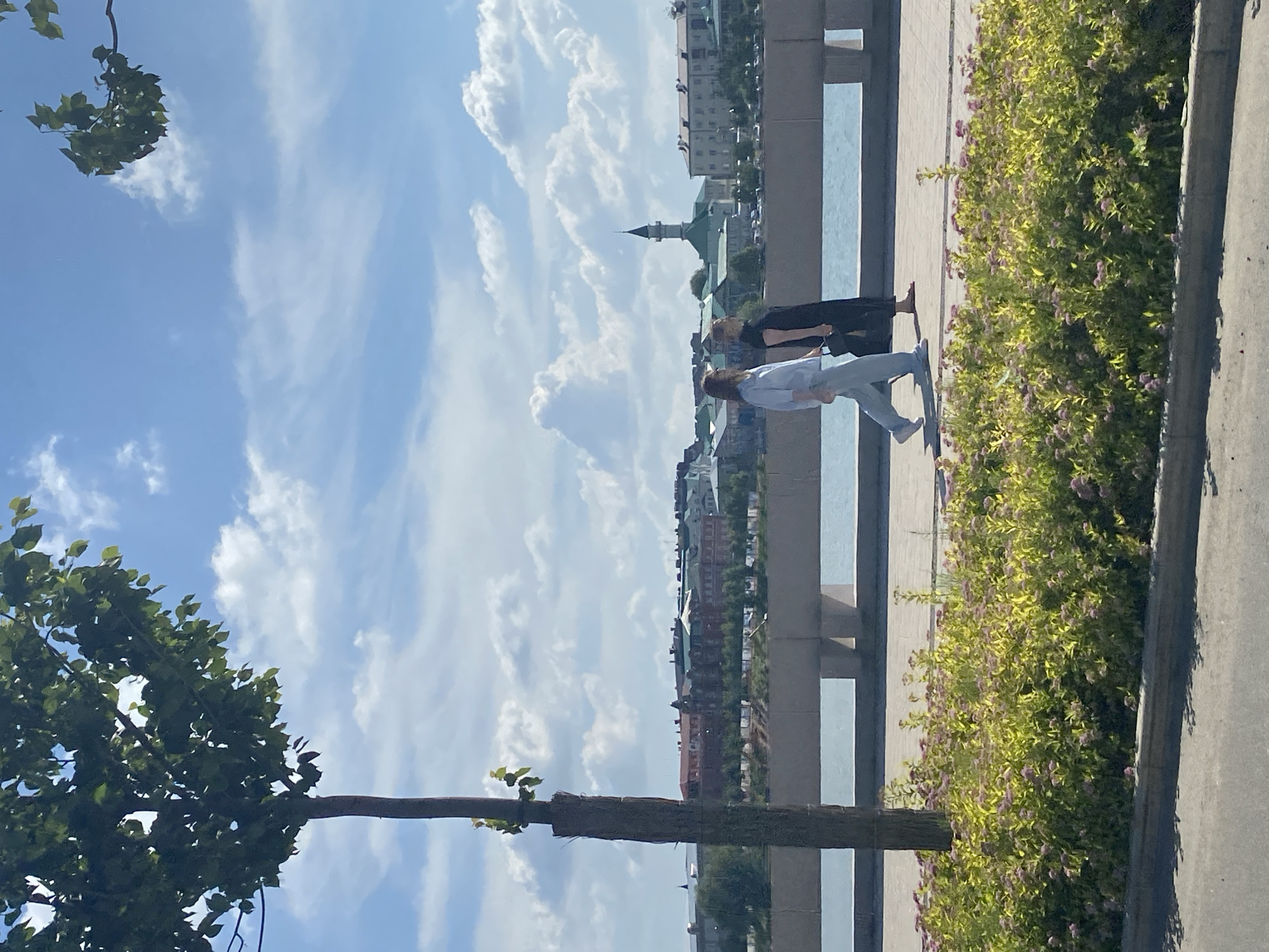 Two individuals are walking along a paved promenade in Kazan, Russia, adjacent to a body of water. The person on the left is wearing light blue attire and appears to be a woman. The person on the right is wearing a black dress and is also a woman. They are walking in the same direction, with the woman in blue slightly ahead.
In the background, a cityscape is visible, characterized by a collection of buildings with various architectural styles. A prominent feature is a minaret with a green spire, suggesting a religious structure, possibly a mosque. The buildings are situated along the waterfront. The water itself is a calm blue, reflecting the sky.
In the foreground, a vibrant green bush with small pink flowers lines the promenade, creating a natural border. To the left of the promenade, a slender tree stands, its trunk wrapped in what appears to be protective material. The sky above is a bright blue, dotted with fluffy white clouds. The overall scene suggests a pleasant day in an urban park or waterfront area.