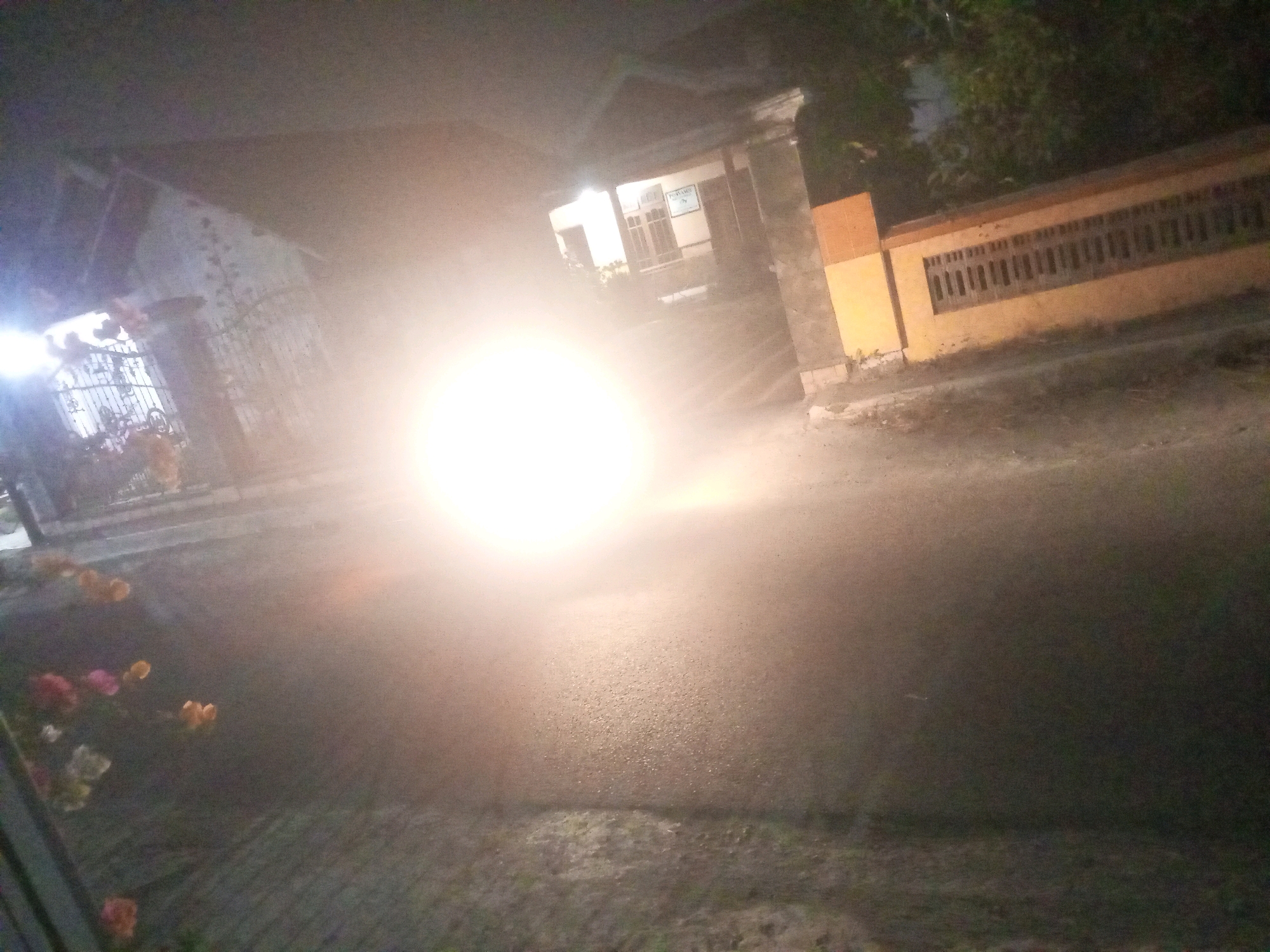 In the city of Ngasem, Indonesia, at night, a street scene is captured with a bright, blinding light originating from the left side of the frame, obscuring much of the foreground and midground. The light appears to be from a vehicle's headlights. To the left, a gate with vertical bars is partially visible, with some foliage or flowers in pink and orange hues in front of it. Behind the gate, a building with a sloping roof and textured walls is present, with a smaller structure attached to its right.
The midground shows a road or paved area, with the intense light reflecting off its surface. Further to the right, a wall with horizontal wooden slats is visible, painted in orange and yellow hues. Behind this wall, dark trees and foliage can be seen, indicating a natural environment. The overall scene suggests a street in a residential or commercial area at night, with the primary element being the overwhelming light source.