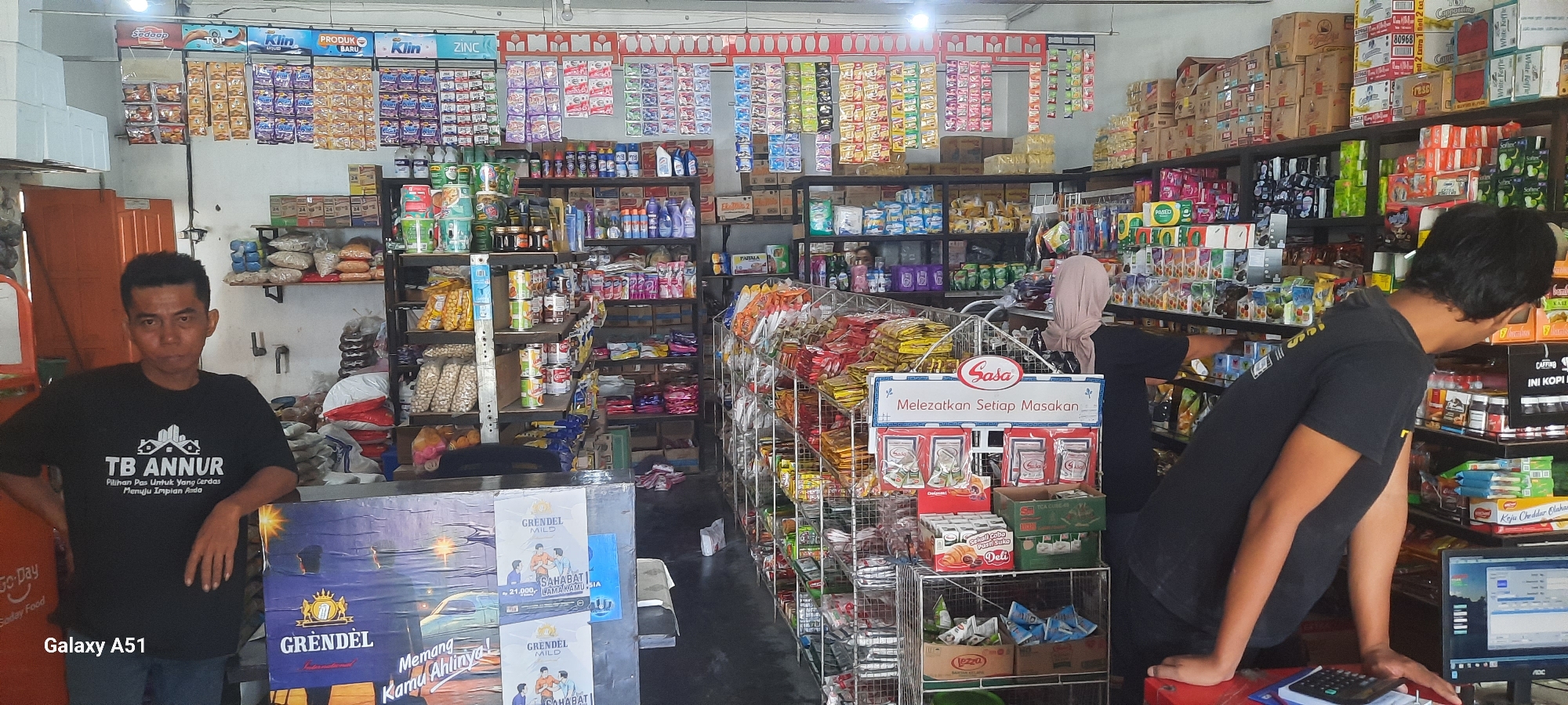 The image depicts the interior of a small convenience store, likely a warung, in Selopuro, Indonesia. A man wearing a black t-shirt with "TB ANNUR" printed on it is standing in the foreground, leaning on a counter. Behind him, shelves are packed with various food products, cleaning supplies, and other household items. To the right, another person, partially visible and wearing a light-colored hijab, is interacting with the merchandise on a display rack. The store appears well-stocked, with products neatly arranged on open shelves and in wire racks. A point-of-sale system with a monitor and calculator is visible on the counter, suggesting active business operations. The overall environment is that of a typical Indonesian neighborhood store.