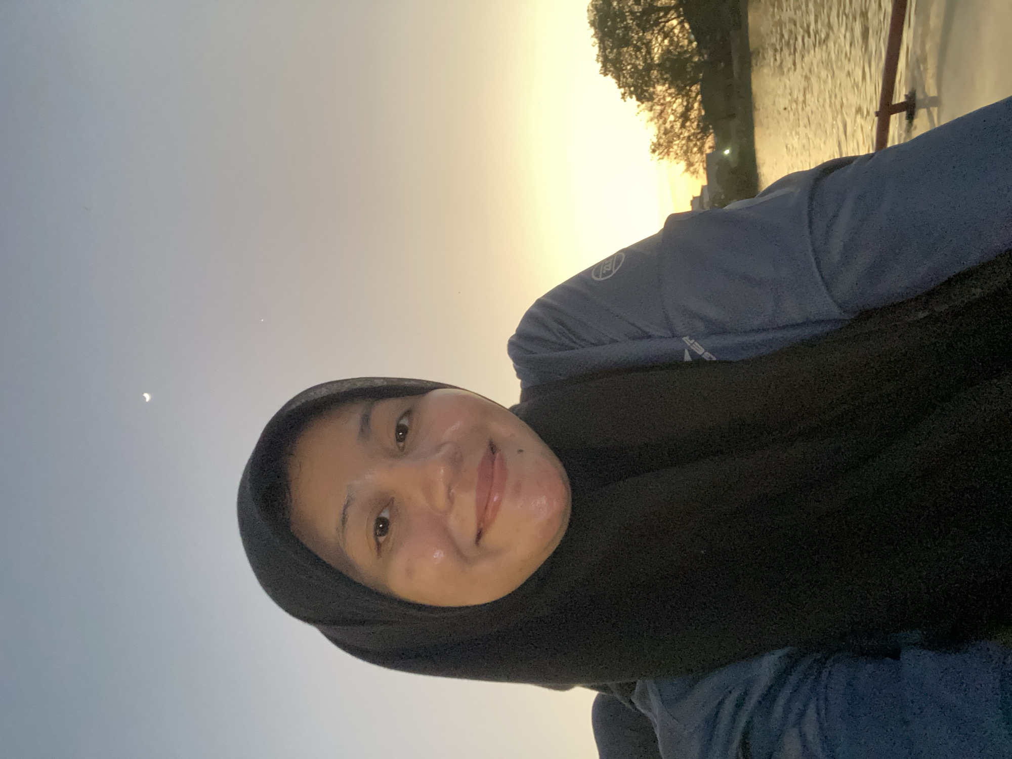 The image shows a woman wearing a black hijab, smiling directly at the camera. She is wearing a blue athletic shirt. The background shows a body of water, possibly a river, with trees and buildings visible on the far bank. The sky is a gradient of soft blues and oranges, indicating a sunset or sunrise. A sliver of the moon is visible in the upper left corner of the sky. The scene appears to be taking place on a boat, as a railing is visible in the foreground. Based on the presence of the river, trees, and general architecture, this scene could be located near the Kuin River in Banjarmasin, Indonesia, a city known for its riverine environment and stilt houses.