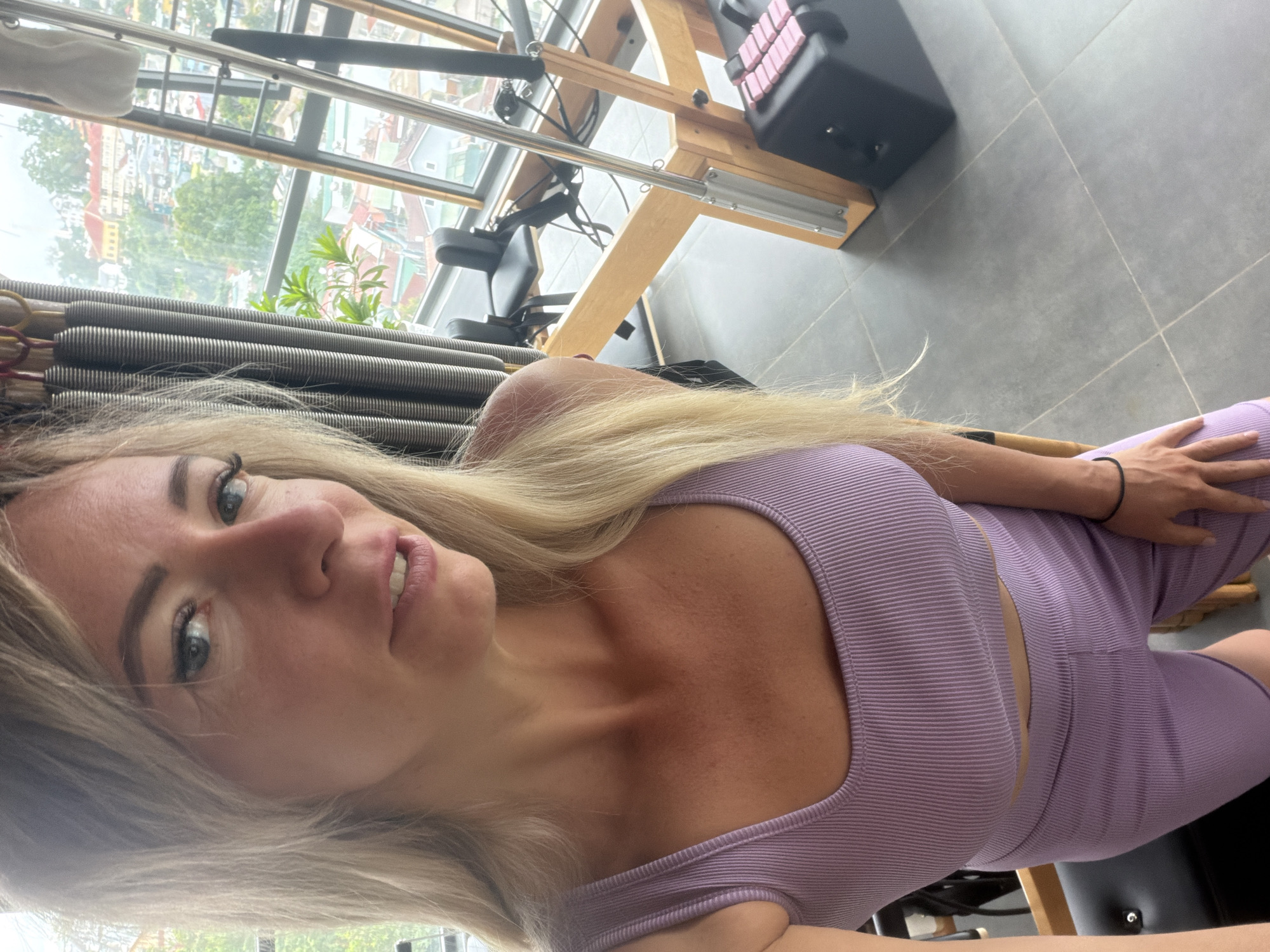 The image shows a woman with blonde hair, wearing a lavender ribbed crop top and shorts, standing in a room with a Pilates reformer in the background. She is facing the camera, looking to the right. The room has large windows overlooking a cityscape with buildings and lush greenery. The environment appears to be an apartment or studio with tiled floors. The cityscape visible through the windows is consistent with the urban landscape of Da Lat, Vietnam, characterized by its hilly terrain and colorful architecture. The Pilates reformer suggests a fitness or wellness setting. The woman's attire and pose indicate she may be preparing for or has just finished a workout.