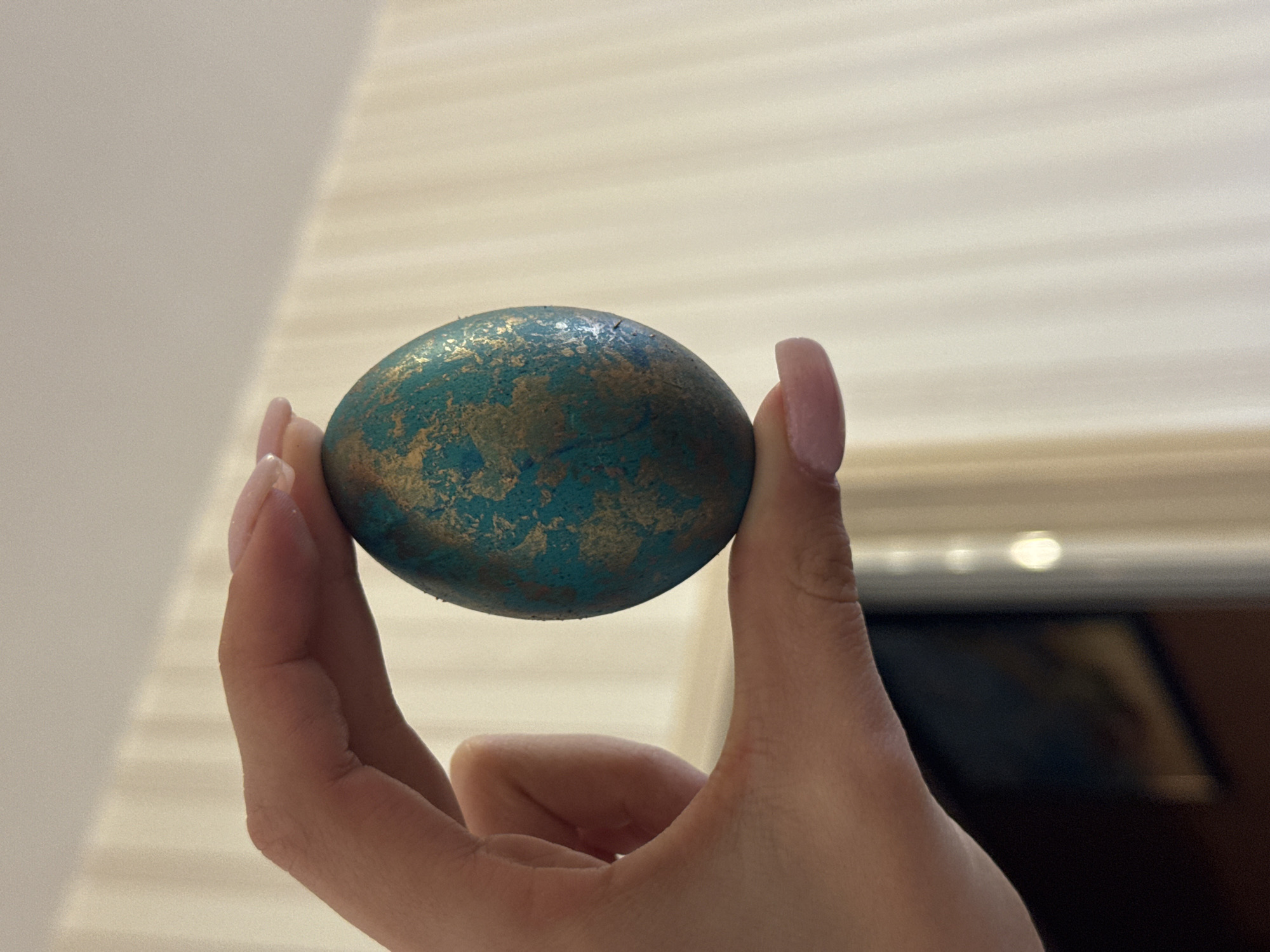 A person with long, light pink acrylic nails holds a round object, likely an Easter egg, between their thumb and forefinger. The egg is painted with an abstract design of teal and gold colors. The background features a white wall with horizontal ridges, and out-of-focus objects suggest an indoor setting. The location in the city of Varna, Bulgaria, is not determinable from the image.