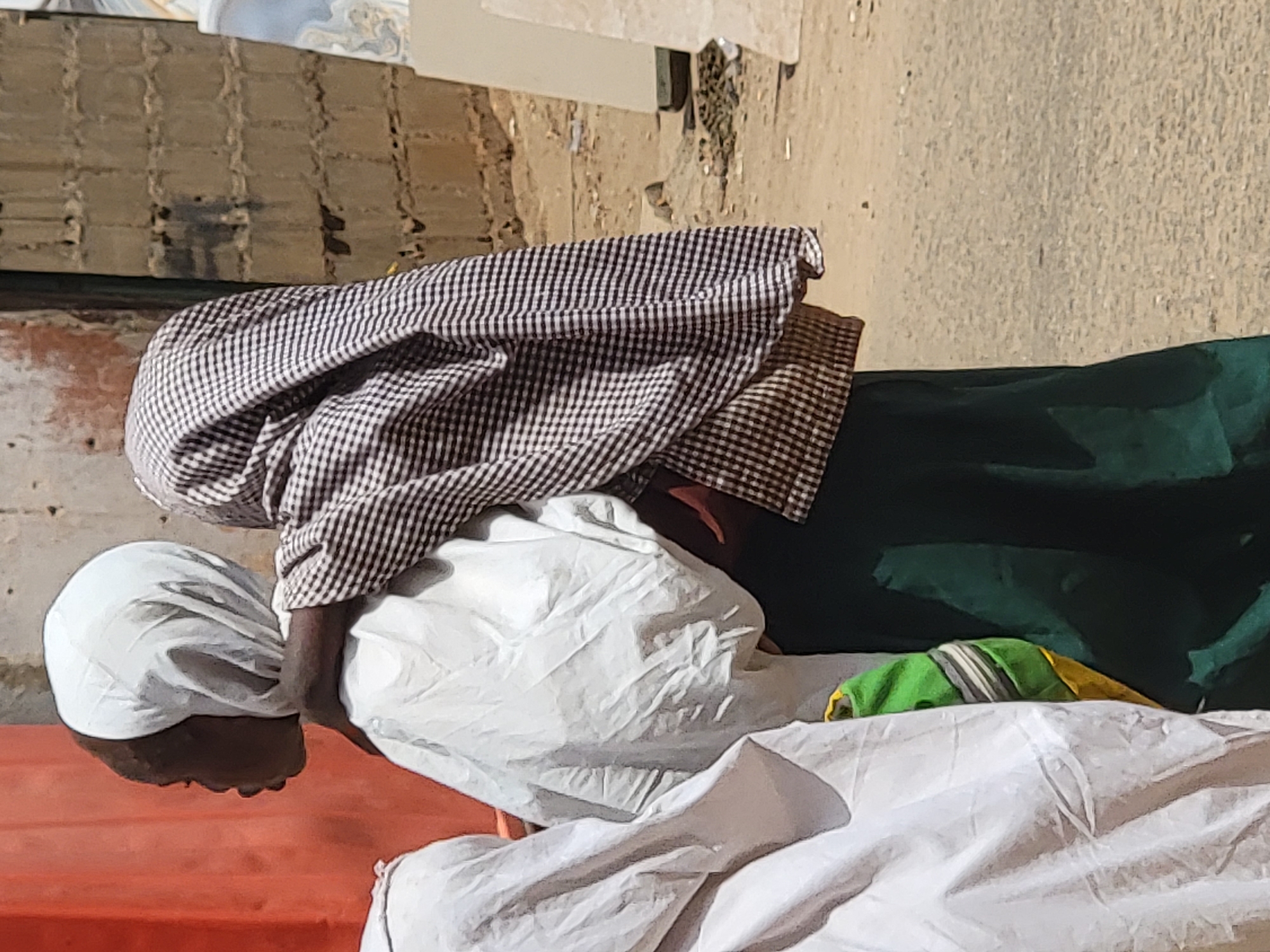 A woman is bent over a white fabric, possibly bedding, on top of something red (perhaps a bed or furniture) in an outdoor setting. The woman is wearing a white head covering, a brown and white checkered garment, and green clothing. The setting appears to be in the city of Maiburiji, Nigeria. The ground is visible in the background and appears to be dirt or concrete. There is a building or wall in the background. The light is bright, suggesting it is daytime.