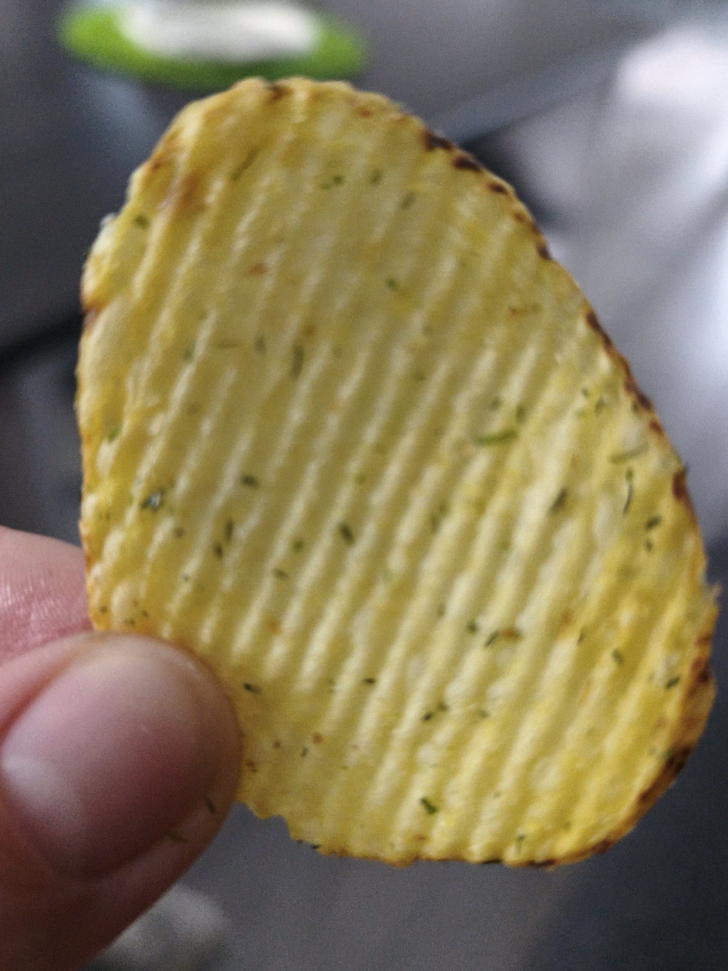 The image shows a close-up of a single potato chip held by a person's thumb and index finger. The potato chip is wavy and yellow, with visible green flecks of seasoning. The background is blurred, with a hint of green and white on the left and a gray surface on the right. There is no discernible location in the city of Brodi, Latvia, visible in the image.