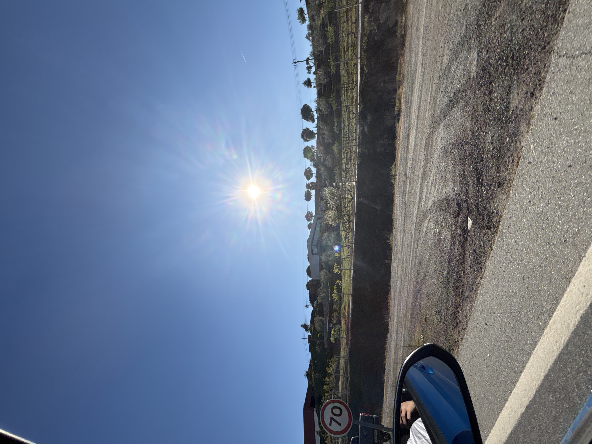 The image depicts a roadside scene in Mirandela, Portugal, captured from inside a moving vehicle. A clear blue sky is visible, with the sun shining brightly, causing lens flare. A rural landscape stretches to the right of the road, featuring trees, shrubs, and possibly vineyards. The road itself is paved and has a white line marking the edge. A speed limit sign reading "70" is partially visible in the lower left, suggesting a speed limit of 70 kilometers per hour. The side mirror of the car is visible in the lower right corner, reflecting the blue exterior of the vehicle and a glimpse of the driver's hand and white sleeve. The general environment indicates a sunny day in a rural area of Mirandela, Portugal.