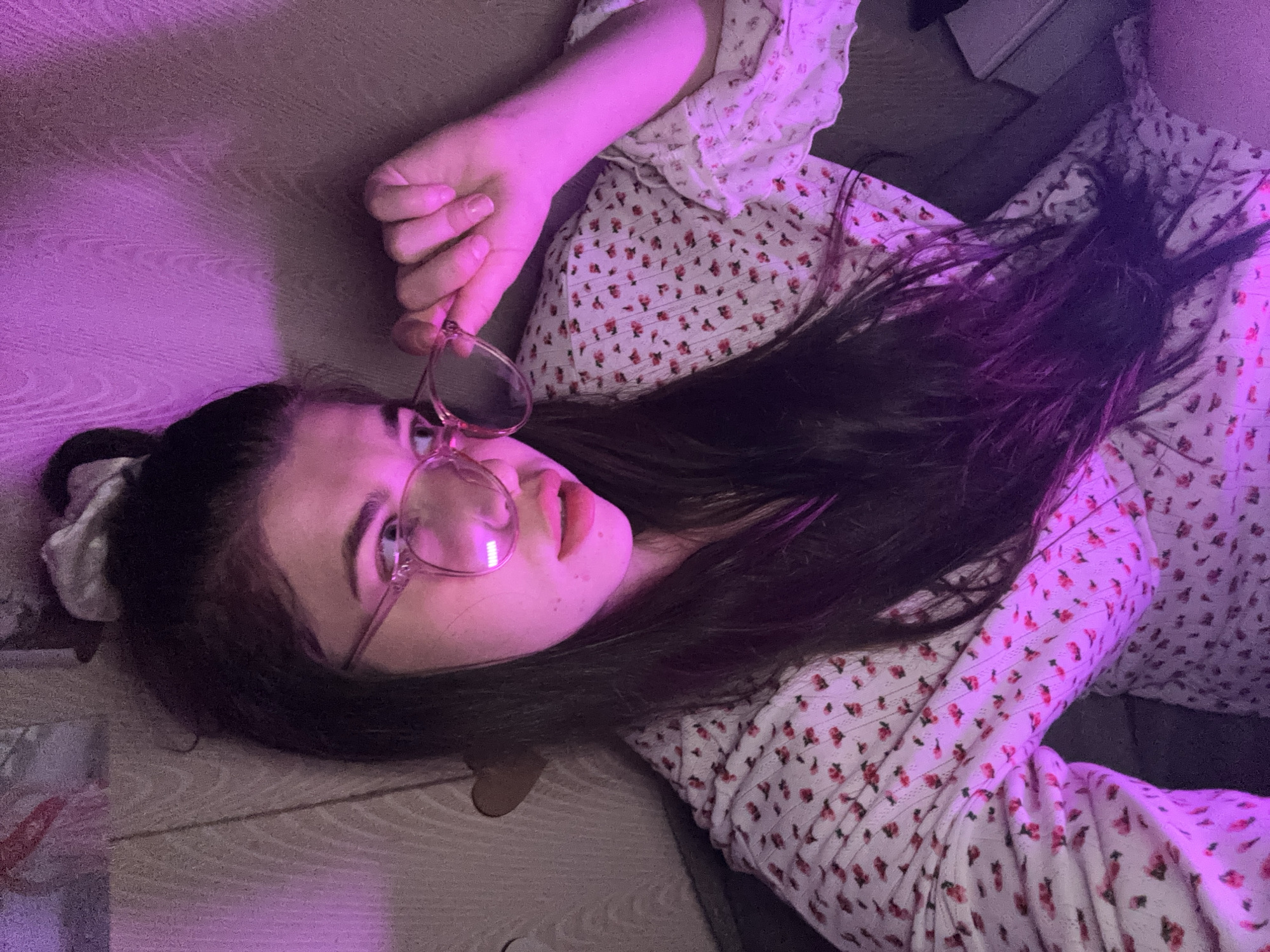 A young woman with dark hair, wearing glasses, lies on what appears to be a wooden floor. She wears a white dress with a small red floral pattern and holds her glasses with her left hand. The lighting in the room has a strong pink hue. The location is not identifiable as the city of Мещёры, Russia based on the provided image.