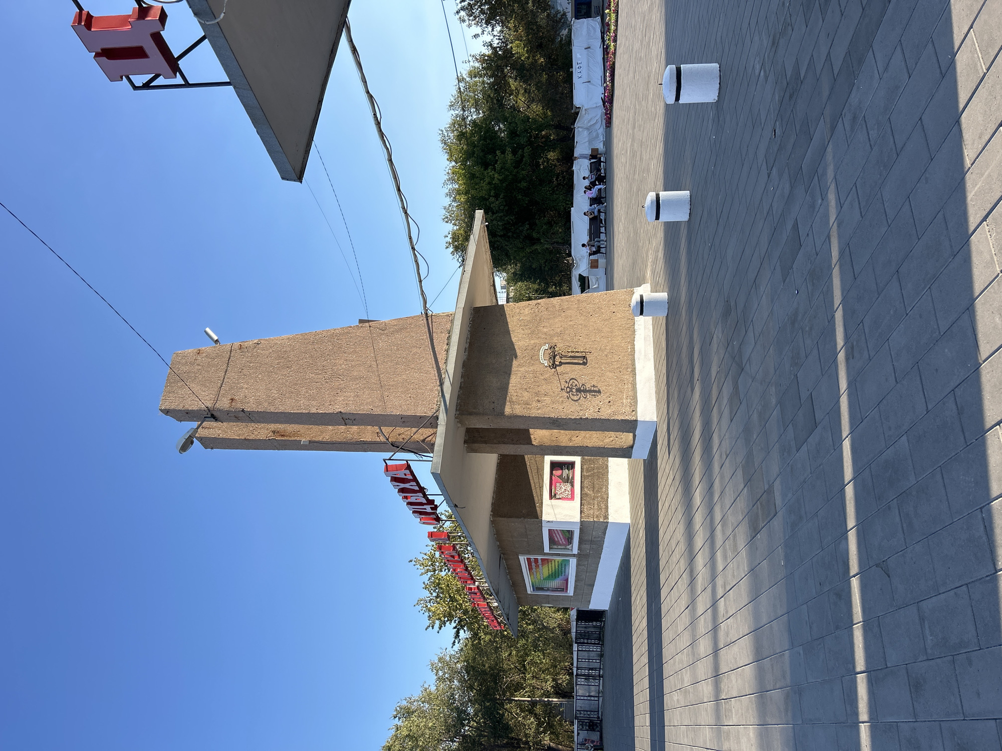 The image captures a sunny day in Orsk, Russia, at what appears to be an outdoor plaza or park area. In the foreground, a paved ground made of gray rectangular tiles is visible, with shadows from a structure casting diagonal lines across it. Three white cylindrical bollards with black tops are spaced out on the pavement.
The central subject of the image is a building with a distinctive architectural style. It features a tall, rough-textured, beige-colored tower-like structure on the left, topped with a simple lamp. Attached to this tower is a cantilevered roof supported by a beige concrete pillar. The building itself is light beige with white accents at its base. Above the entrance area, prominent red Cyrillic lettering spells out "ПАРК ОТДЫХА" (PARK OF REST). Below the sign, there are two rectangular windows, one displaying colorful information, and the other a QR code. A decorative element resembling a key is etched onto the concrete pillar.
To the right of the main structure, a portion of another building or canopy is visible, also with a concrete roof. In the background, beyond the paved area, a row of trees with green foliage is present. Further back, several people are seen sitting on benches, with white construction barriers or temporary fencing behind them. A sign on the white barriers reads "КАФЕ" (CAFE). The sky is a clear, bright blue, indicating good weather.