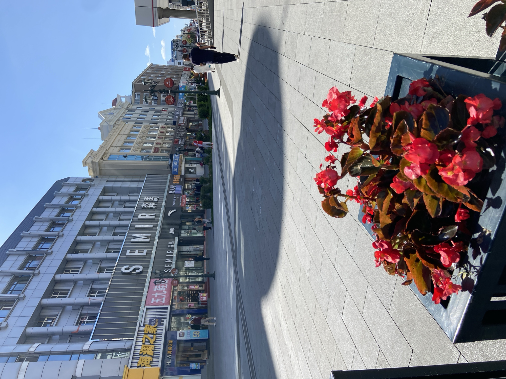 The image depicts a bright, sunny day on a pedestrian street in Xing'an Subdistrict, China. In the foreground, a dark planter box holds vibrant red flowers with dark green leaves. The street is paved with large, light-colored rectangular tiles, casting long shadows from an unseen object on the left. A single person, dressed in dark clothing, walks away from the camera down the street on the right side of the frame. Several other pedestrians are visible further down the street, walking or standing near shop entrances. The buildings lining the street are multi-story structures with a mix of modern and slightly older architectural styles, featuring large glass windows and various signage. Notable signs visible include "SEMIR" and "ANTA". The sky is clear blue.