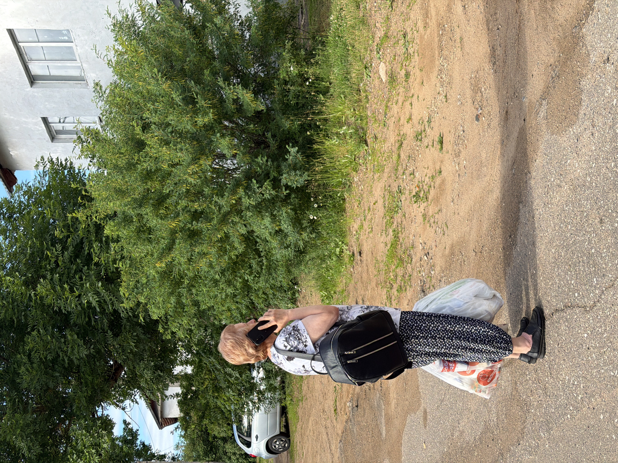 This media file depicts an outdoor scene in Moshenskoe, Russia, likely during daytime given the shadows. A woman with short, light-colored hair is the primary subject. She is wearing a black and white floral print top, a black and white patterned skirt, and dark shoes with pink socks. She is holding a black smartphone to her ear, suggesting she is engaged in a phone conversation. A black backpack with zippers is slung over her shoulders, and she is carrying two large plastic bags, one white and one with red lettering.
The woman is standing on a paved surface, possibly a sidewalk or road, which appears slightly wet. To her left, there is a dirt and grass area with a large, leafy green tree and some smaller plants and white flowers. Behind the vegetation, a pale grey building with at least two windows is visible. Further in the background, a light-colored car is parked near the building, and more trees and foliage can be seen.
The overall impression is a casual, everyday moment. The lighting suggests it is a bright day, possibly late morning or afternoon. The setting appears to be a residential or semi-rural area, given the presence of a building, car, and natural vegetation. There is no strong indication of emotion, but the woman's posture suggests she is occupied with her phone call. The visible text on one of the bags is partially obscured, but the word "ОРЕО" is discernible in red lettering.