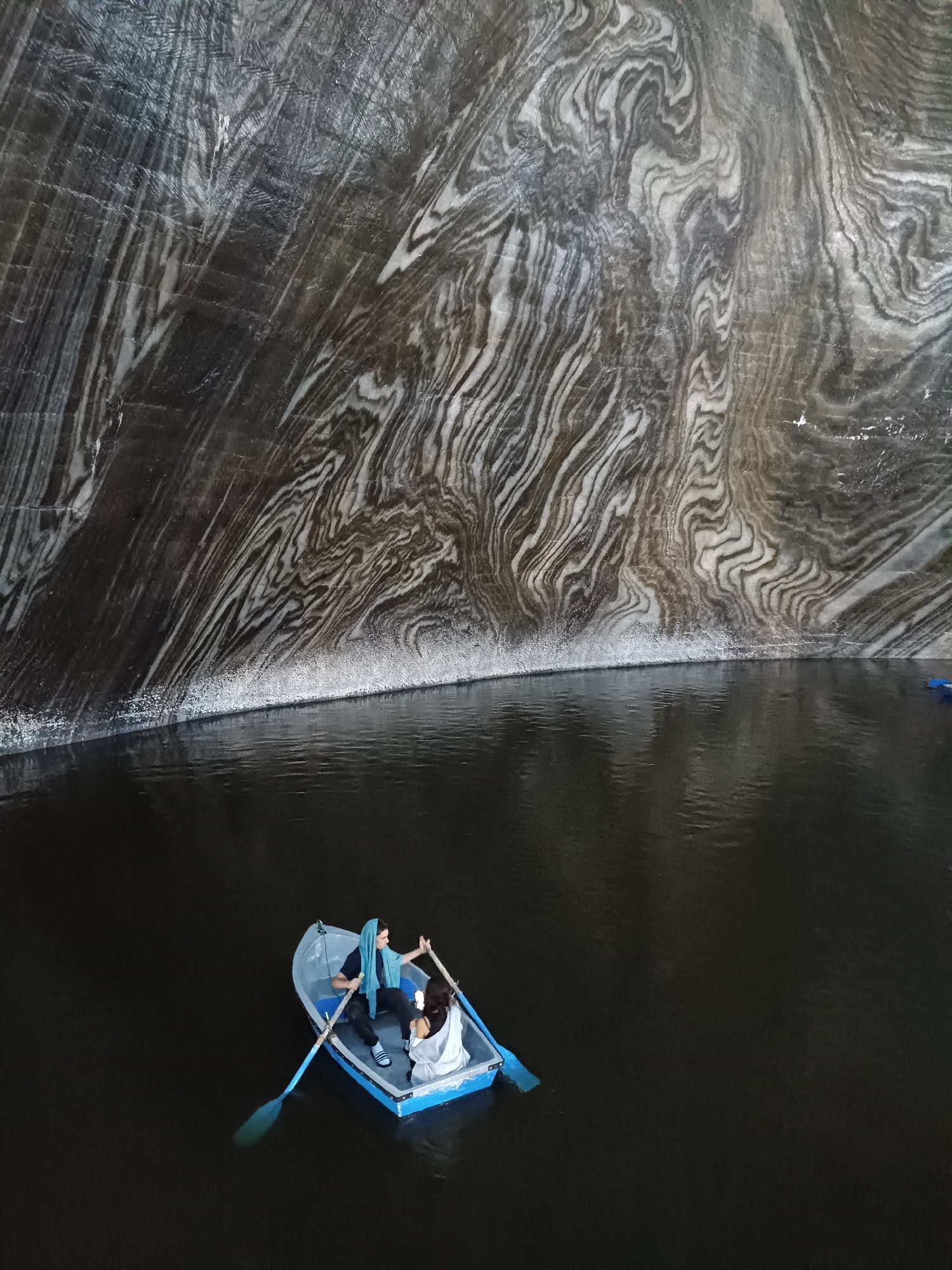 The image depicts an underground salt mine in Turda, Romania, specifically the Salina Turda. The foreground features a dark, still body of water, likely a lake formed within the mine. A small blue and white rowboat is in the water, carrying two individuals. One person, wearing a blue headscarf, is seated and holding oars, appearing to be rowing. The second person, wearing white, is seated in the back of the boat. The background is dominated by the massive, textured walls of the salt mine, characterized by dramatic striations of dark grey and white salt deposits. These geological formations create a surreal and cavernous atmosphere. The lighting is dim, highlighting the textures of the salt walls and the reflections on the water.