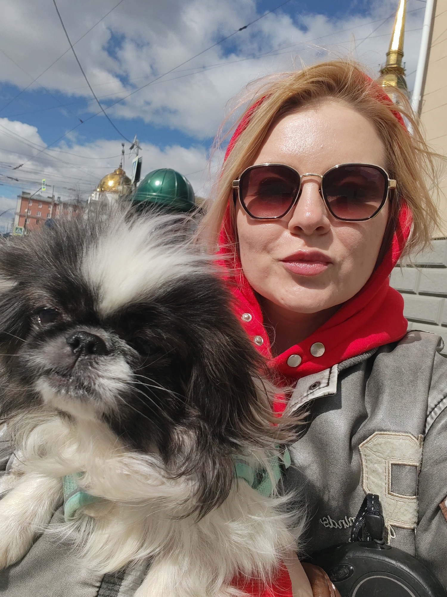 A woman with blonde hair and sunglasses holds a black and white Pekingese dog in her arms. She wears a red hoodie underneath a grey leather jacket. The background includes a blue sky with clouds, building rooftops with gold and green domes, and street elements. The presence of domes characteristic of Russian Orthodox churches suggests the city could be Izhevsk, Russia.